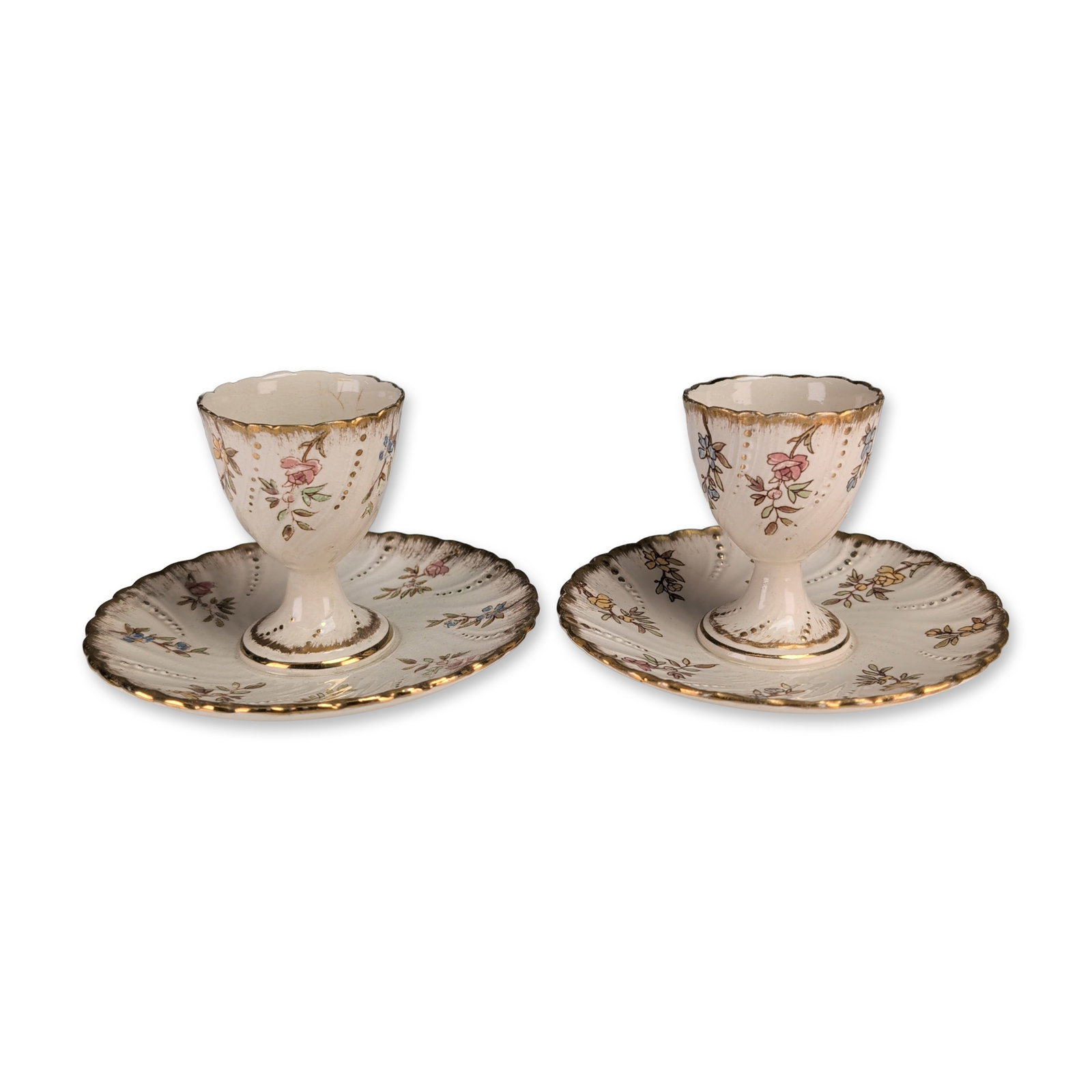 Pair of Sarreguemines Louis XV French Earthenware Egg Cups: Pair of French earthenware egg cups by Sarreguemines in the Louis XV pattern. Each pedestal-form cup is permanently joined to a wide, lobed integral saucer with a scalloped edge. The cream-colored bod