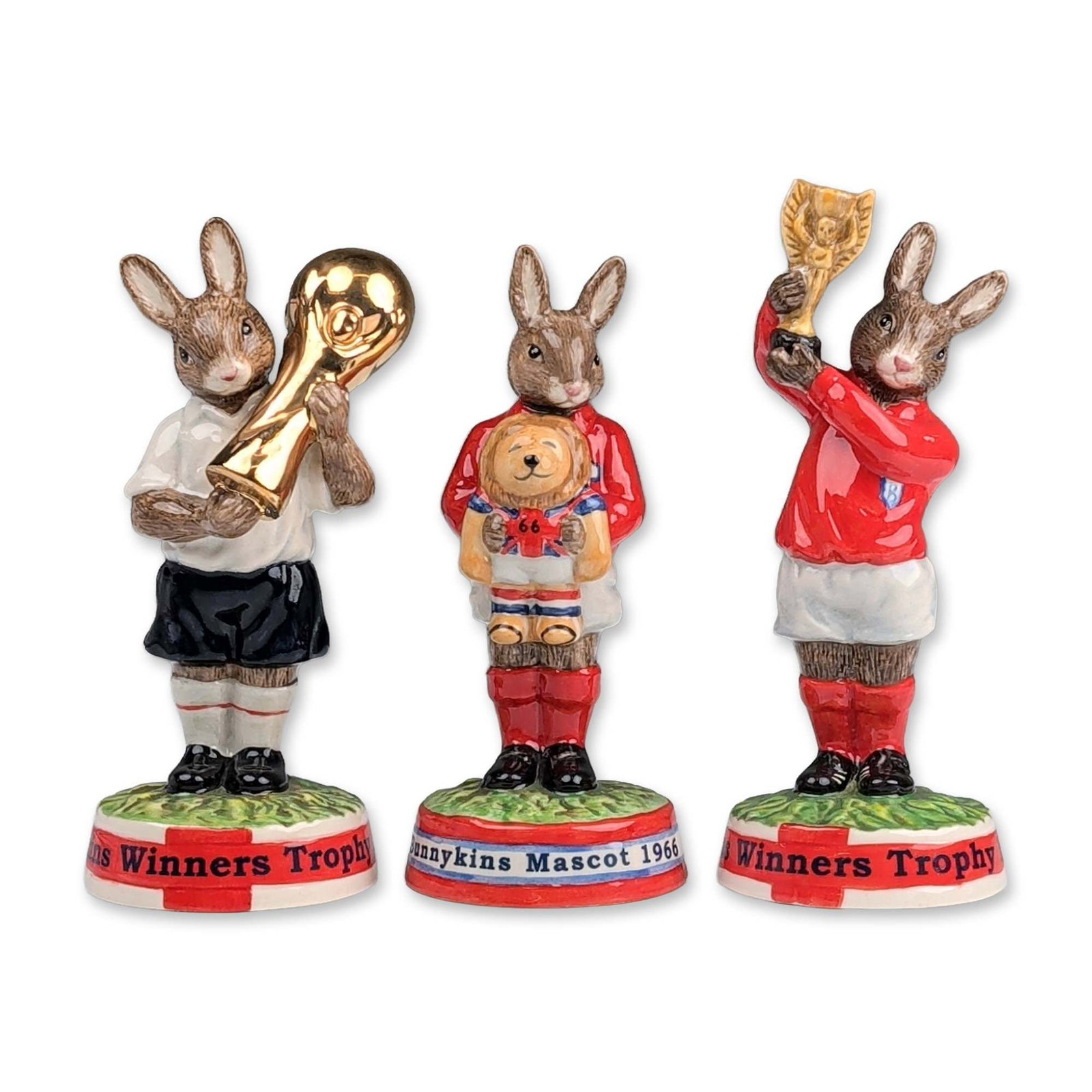 Trio of Royal Doulton Bunnykins World Cup Figurines: Royal Doulton. Trio of hand-painted porcelain Bunnykins figurines themed around World Cup soccer. The collection includes Winners Trophy 1966 (DB 410) with a rabbit in a red kit holding the Jules Rime