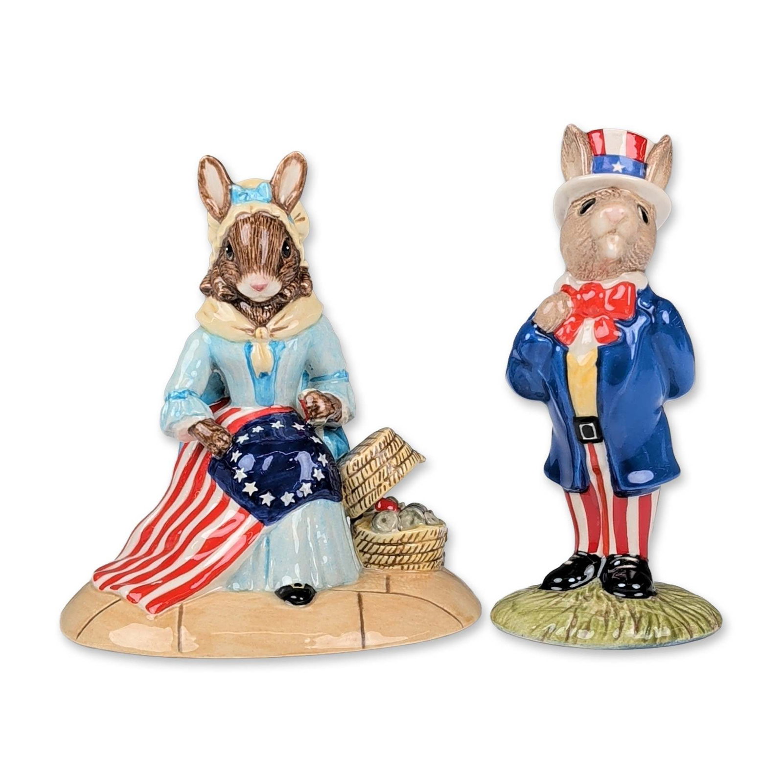 2pc Royal Doulton Bunnykins American Patriotic Figurines: Royal Doulton, England. Pair of hand-painted porcelain figurines from the Bunnykins series featuring American patriotic themes. One figurine depicts Uncle Sam dressed in a blue frock coat, yellow wais