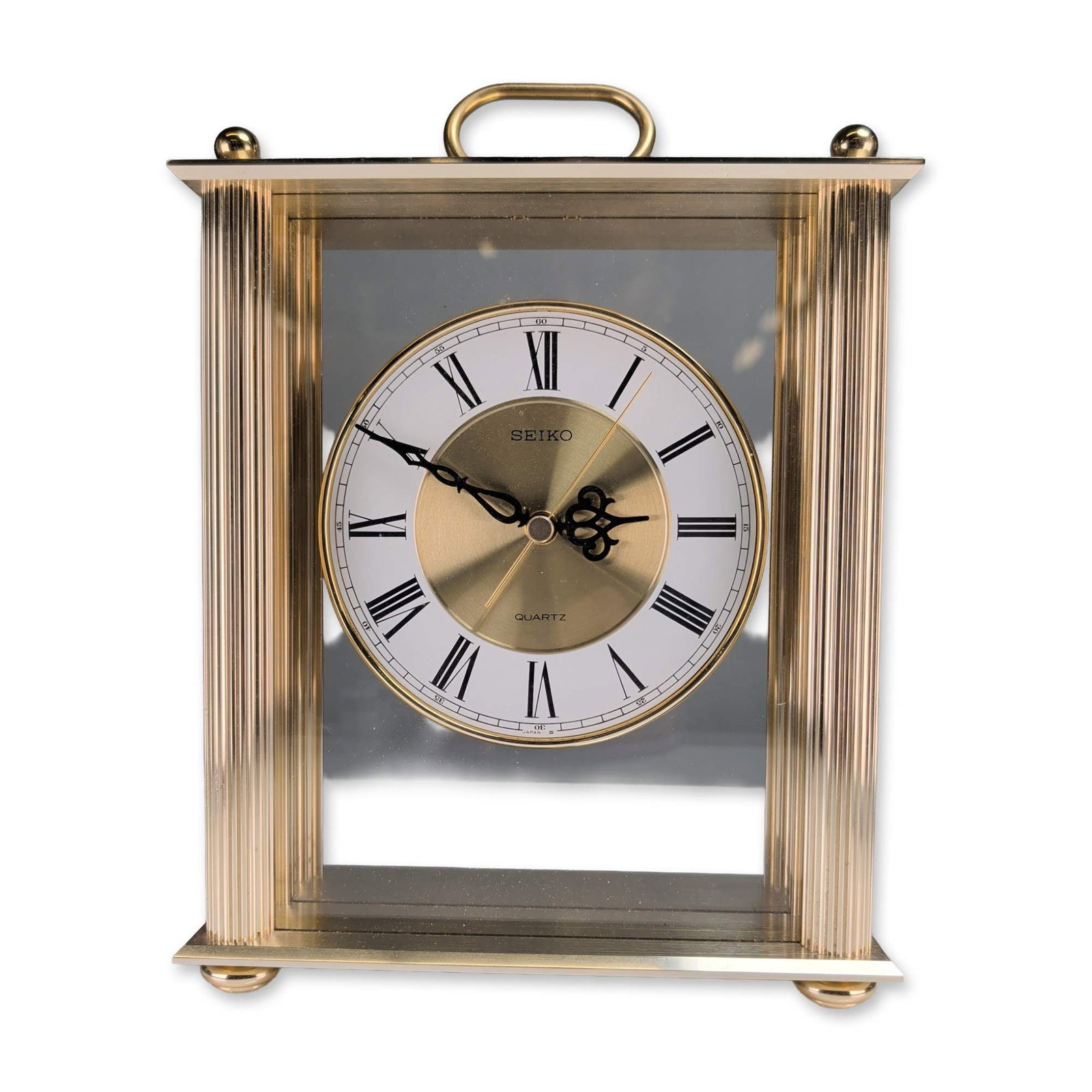 Seiko Gold Tone Quartz Carriage Clock: Seiko carriage style mantel clock in a gold tone metal and glass case with fluted corner pillars and a swinging top handle. The circular dial is configured with a white chapter ring, black Roman numer