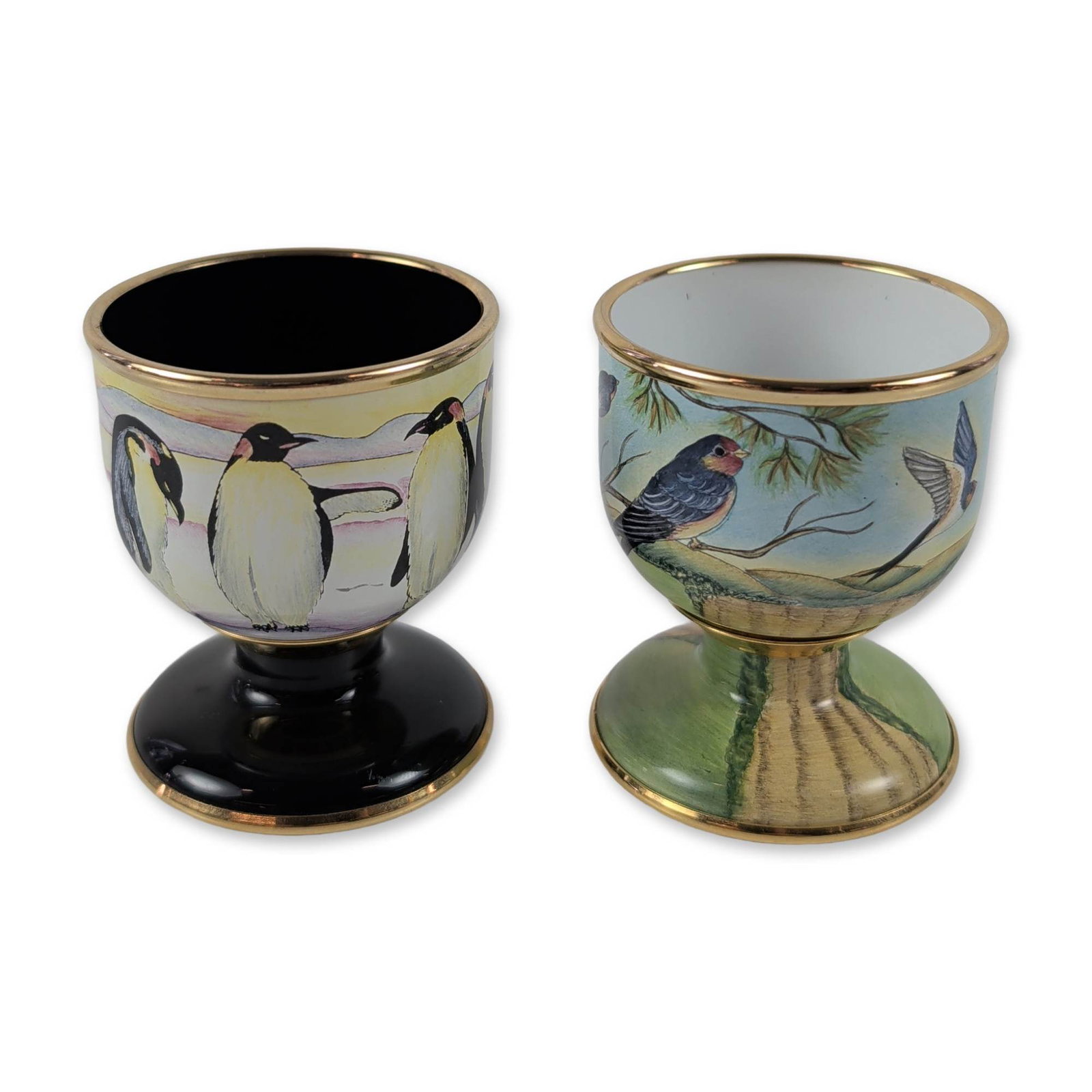 Pair of Moorcroft Enamel Bird Egg Cups: Moorcroft Enamels. Pair of hand-painted enamel pedestal egg cups featuring bird motifs and gilded highlights. One cup is decorated with swallows among pine branches with a green landscape pedestal and