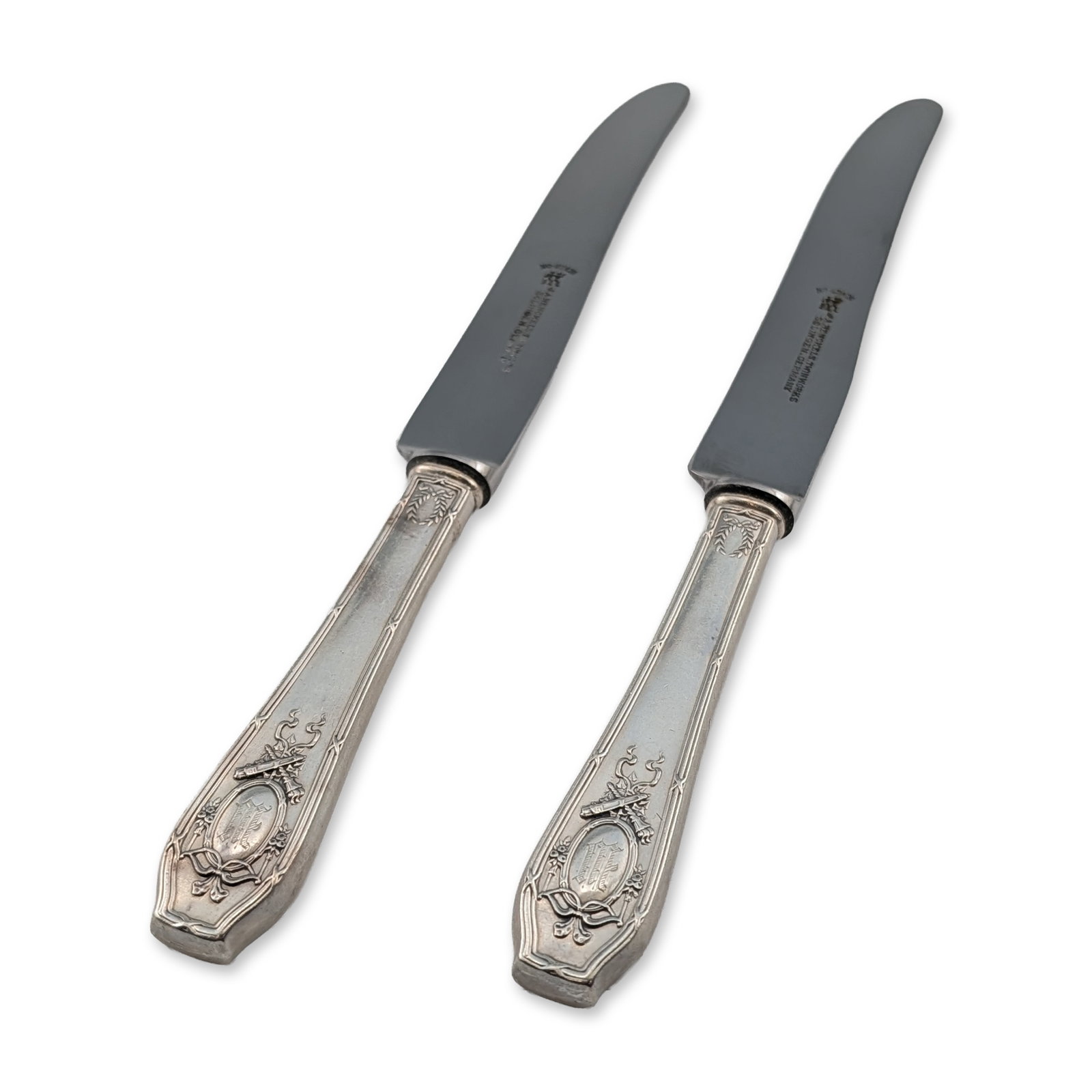 2pc R. Wallace & Sons Sterling Silver Dinner Knives: Pair of dinner knives featuring weighted sterling silver handles in a Neoclassical design with laurel wreaths, ribbons, and foliate scrolls. Each handle includes a central oval cartouche engraved with
