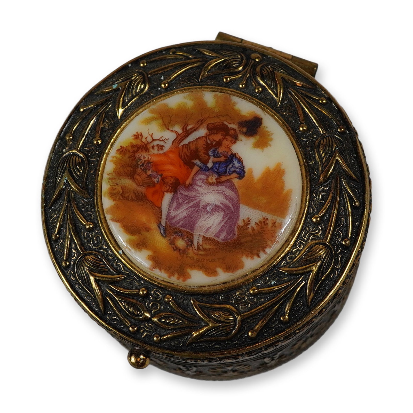 West German Gold Toned Jewelry Treasure Box with Hand Painted Scene Plaque Signed Fragonard: A circular jewelry treasure box, also suitable for use as a pill box, manufactured in West Germany. The piece is constructed from gold-toned metal with an ornate embossed design. The sides of the box 
