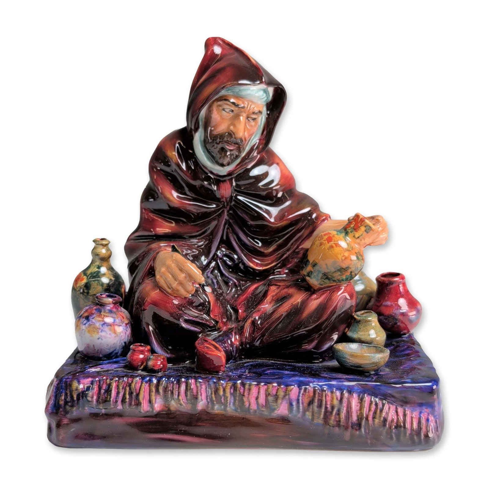 Royal Doulton Bone China Figurine, The Potter HN1493: Designed by Charles Noke. The seated figure is depicted in a heavy hooded robe and turban, surrounded by various small pots finished in multi-colored glazes including green, blue, and yellow. The main
