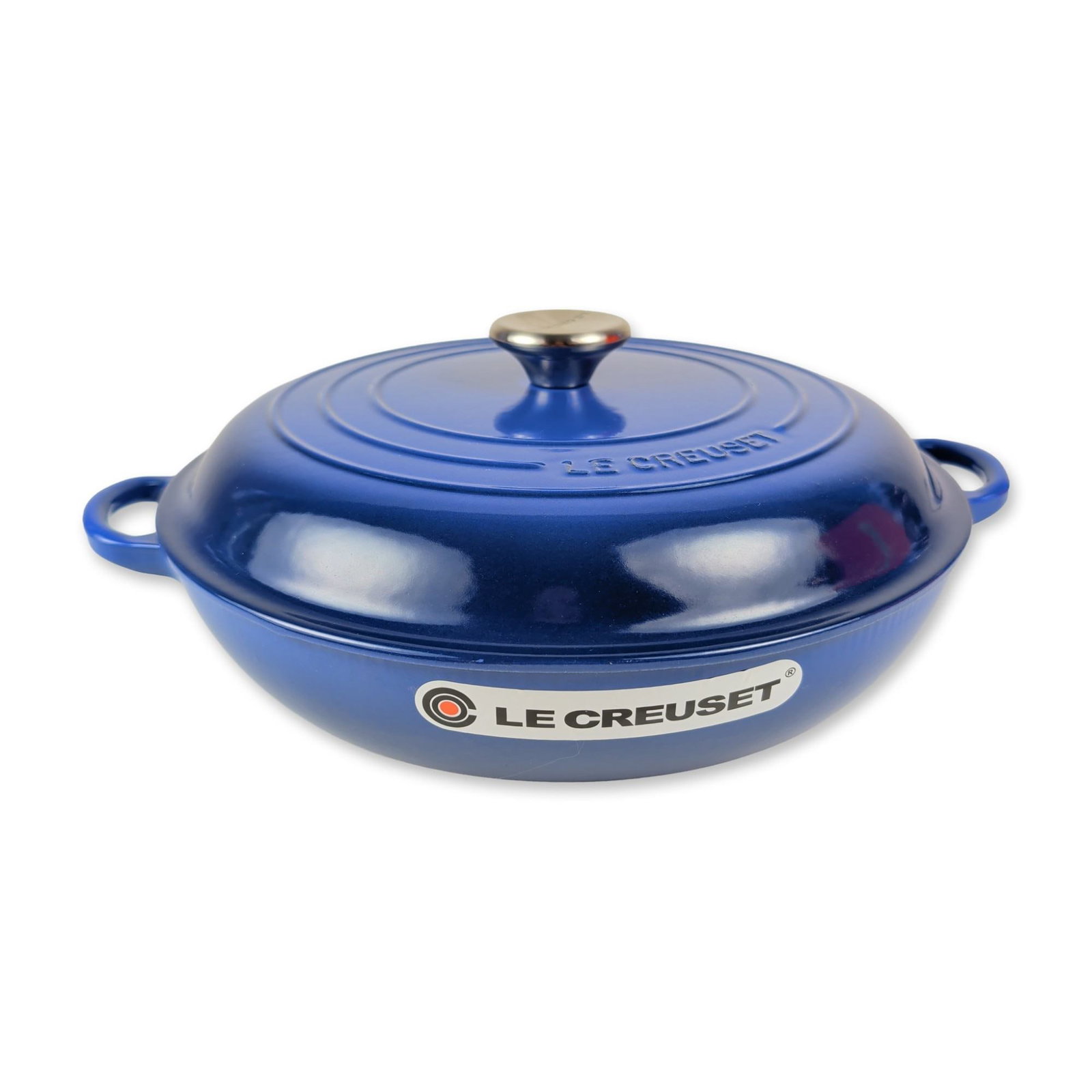 Le Creuset Cobalt Blue Enameled Cast Iron Shallow Braiser: Cobalt blue exterior finish and a cream-colored enamel interior. The pot is designed with a rounded base and two integrated loop side handles. The matching domed lid features the signature three conce