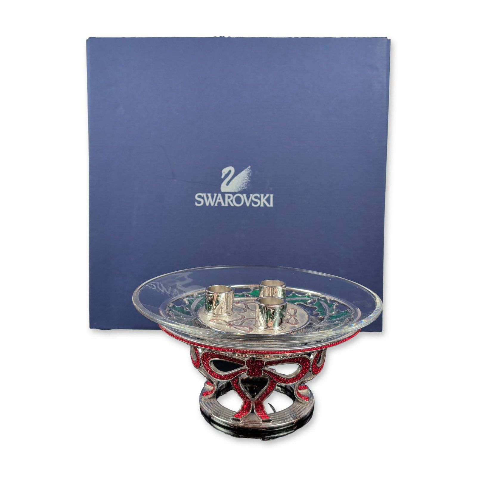 Swarovski Holiday Crystal and Metal Candleholder: Swarovski three-piece holiday candleholder set in clear faceted crystal and silver-tone metal. The assembly consists of a pedestal base decorated with red pave crystal bows, a shallow clear crystal bo