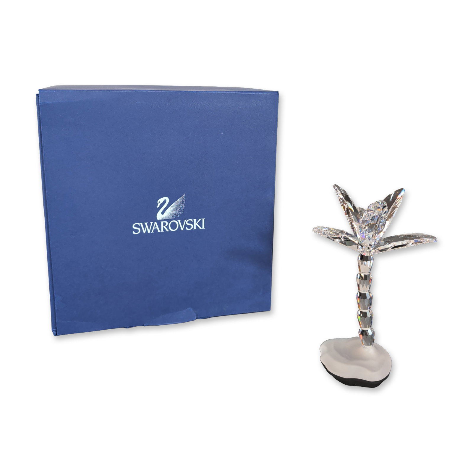 Swarovski Crystal Palm Tree Figurine: The piece features a crown of clear faceted crystal fronds atop a trunk composed of stacked faceted segments. The tree is mounted to a frosted crystal base with a naturalistic, undulated edge. The und