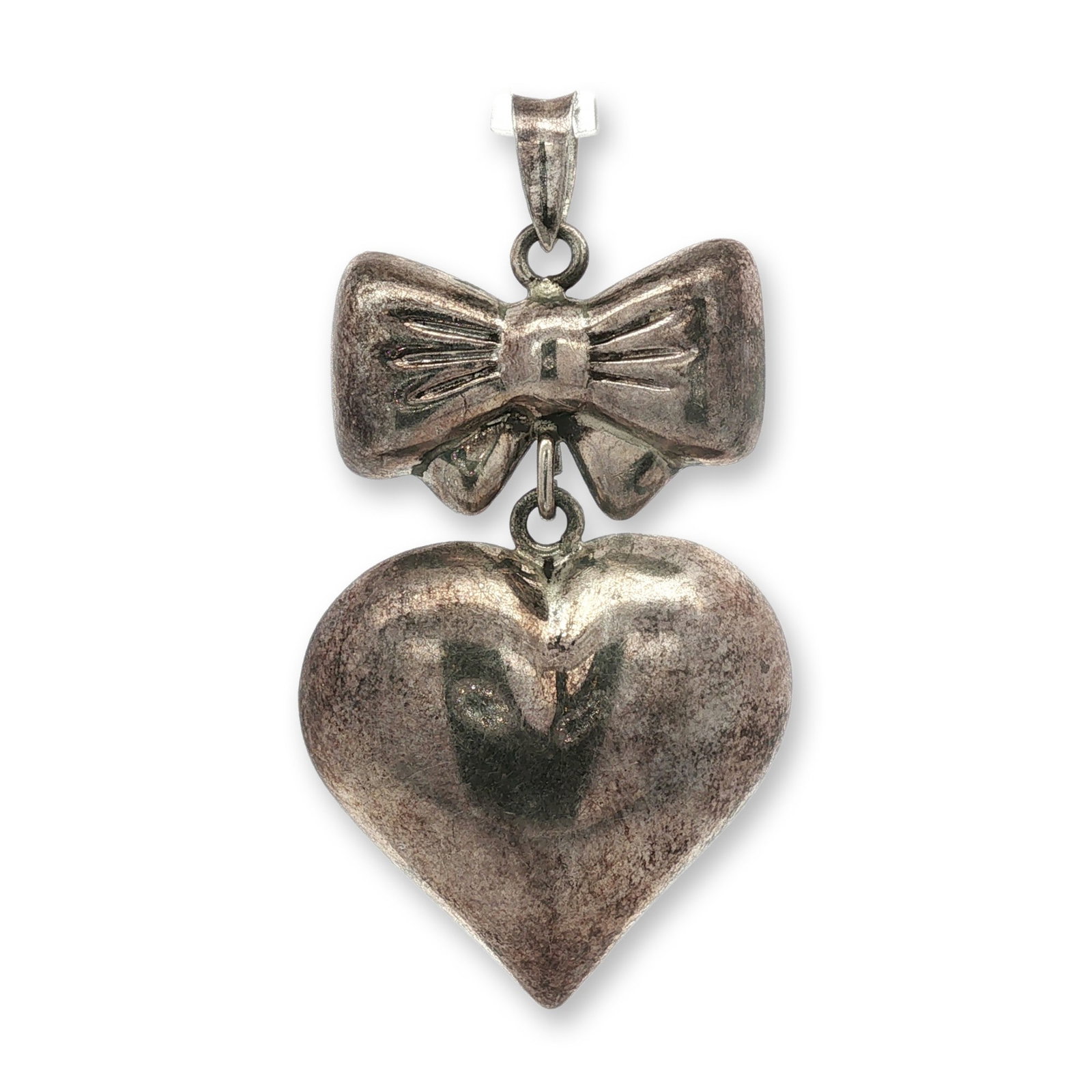 Sterling Silver Bow and Puffy Heart Pendant: Sterling silver pendant featuring a stylized bow with incised linear texture and central knot, from which a three-dimensional puffy heart is suspended. The piece is constructed with a fixed loop and t