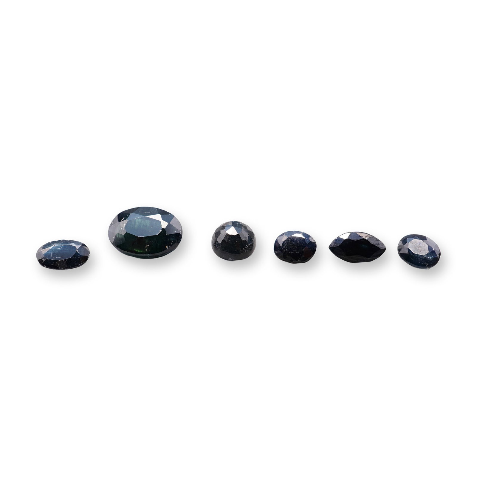 6pc Loose Faceted Deep Blue Sapphires: Collection of six loose faceted gemstones, appearing to be sapphires, displaying a deep blue hue. The group includes a variety of cuts including marquise, round, and oval. The gemstones exhibits