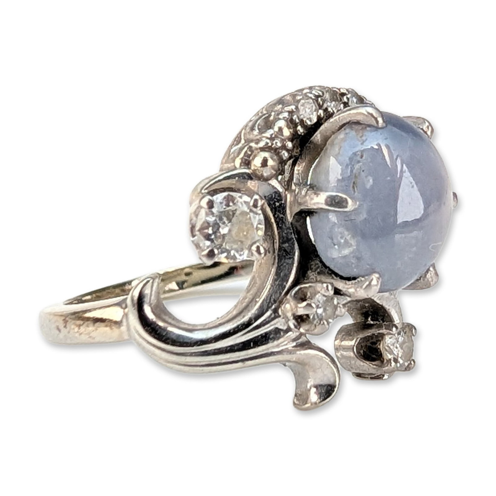 14K White Gold Star Sapphire and Diamond Cocktail Ring: Mid-century cocktail ring in 14k white gold, featuring a center oval cabochon-cut light grayish-blue star sapphire. The star sapphire is held in a six-prong setting and exhibits visible asterism. The 