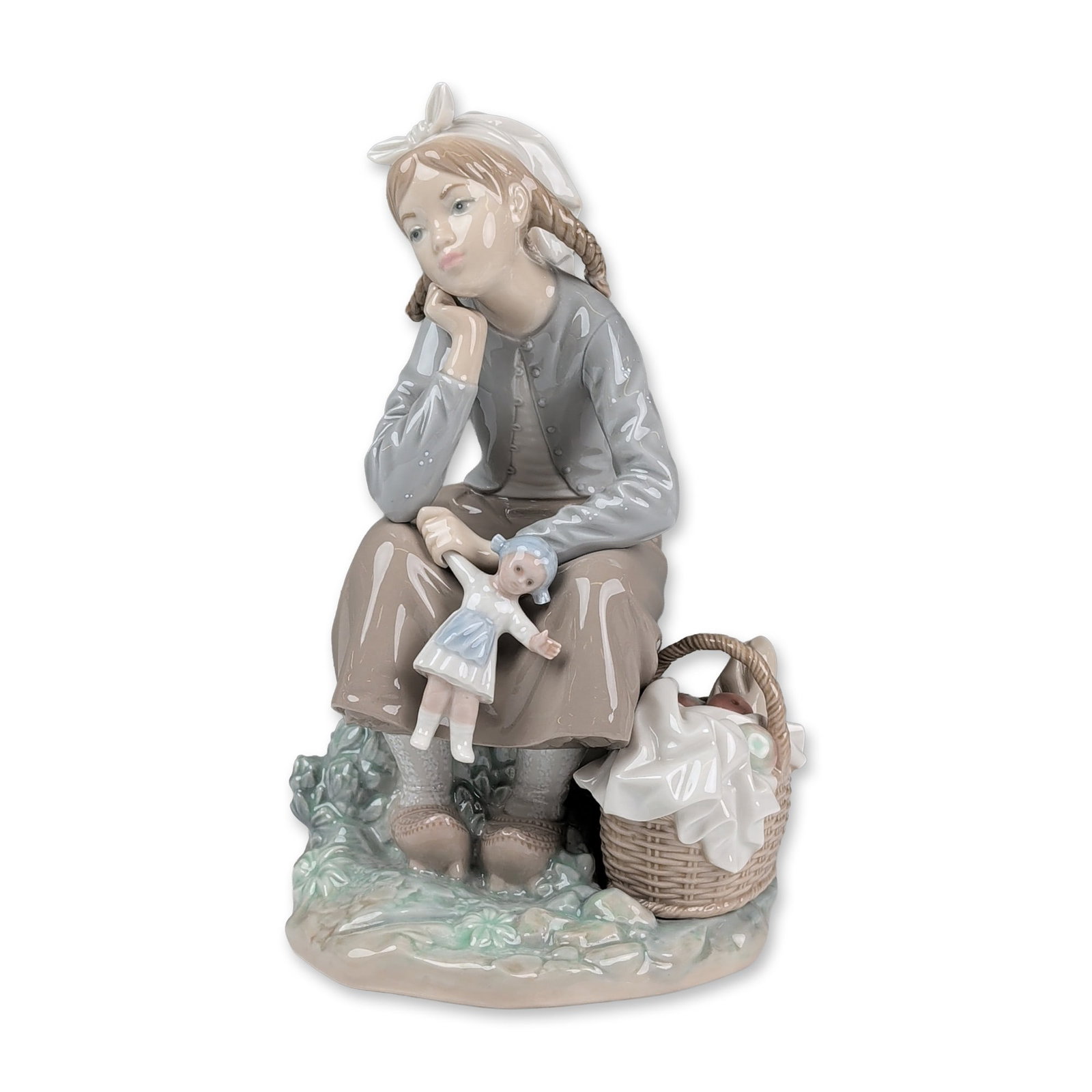 Lladro Porcelain Figurine, Girl with Doll 01001211: The sculpture depicts a young girl wearing a bonnet and long skirt seated on a rocky outcrop. She holds a small doll in her lap, with a wicker basket of apples and a tied parcel at her feet. The piece
