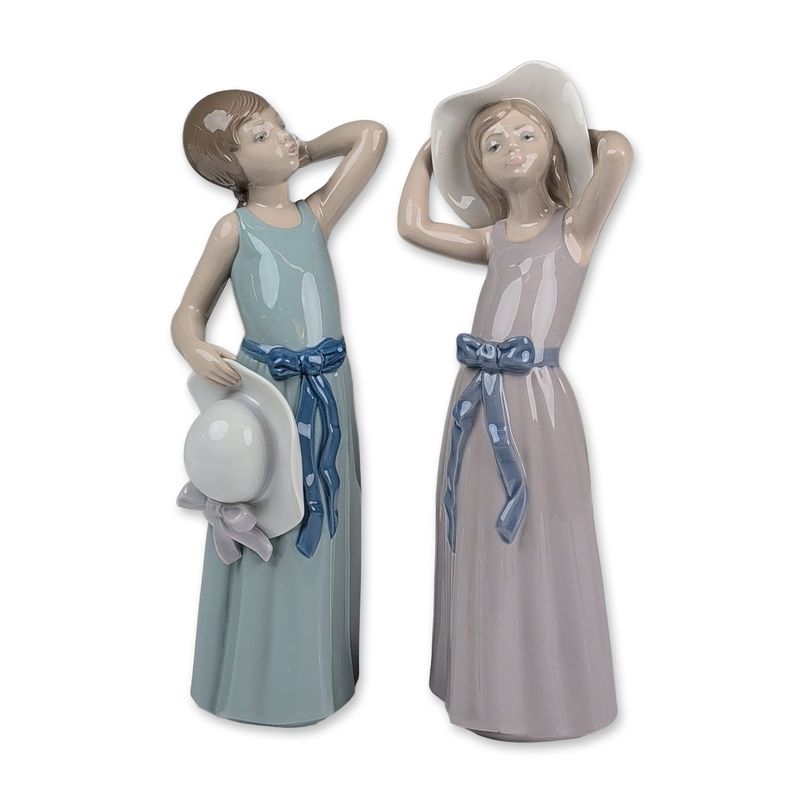 2pc Lladro Glazed Porcelain Figurines, Prissy 01005010 & Coy 01005011: Two glazed porcelain figurines, each figure depicts a young woman in a floor-length dress accented with a blue sash and bow. Coy figurine (5011) is modeled in a lavender-gray dress holding a wide-brim