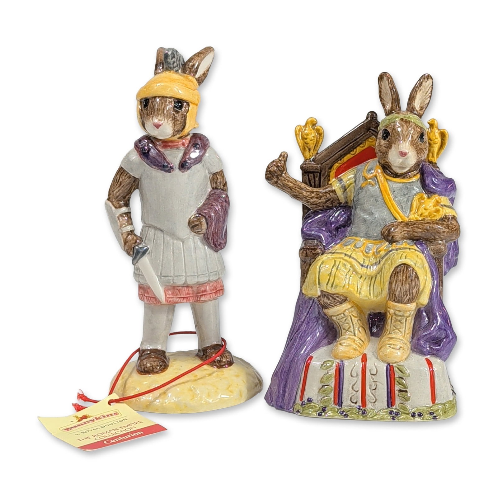 2pc Royal Doulton Bunnykins Roman Empire Figurines: Figurines from the Bunnykins Roman Empire Collection. This lot includes Emperor Bunnykins, model DB312, depicted seated on an ornate throne in a purple toga, and Centurion Bunnykins, model DB 294, dep