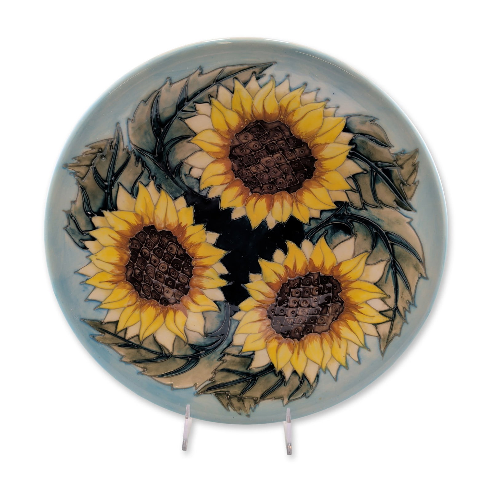 Moorcroft Pottery Sunflower Cabinet Plate: The circular form features hand-painted tube-lined decoration depicting three large sunflower blossoms in yellow and ochre hues with dark brown centers, set among stylized green leaves on a pale blue 