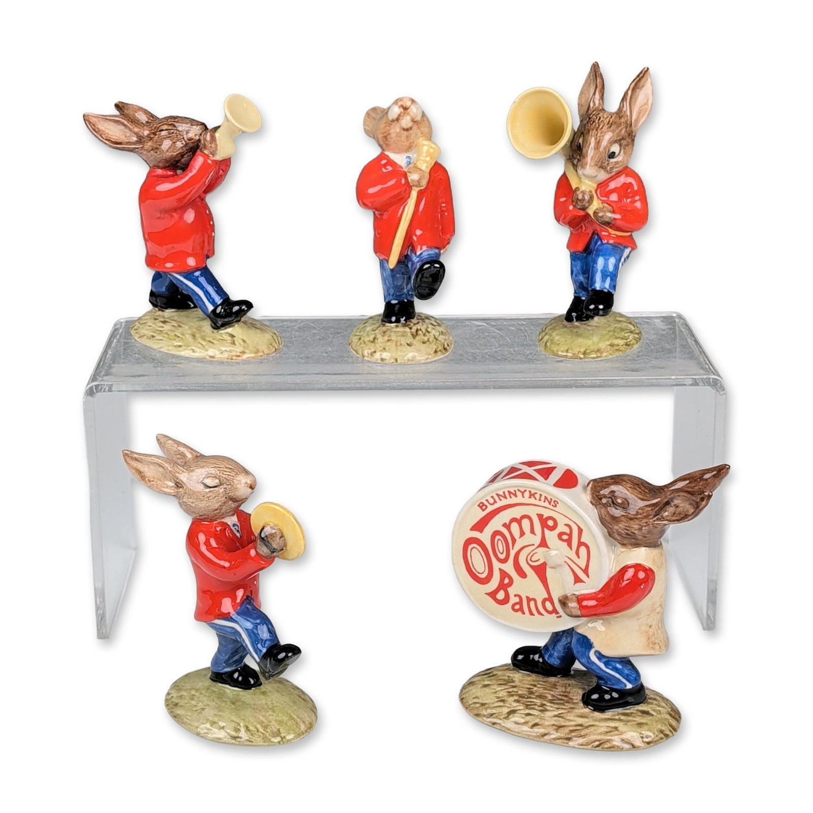 5pc Royal Doulton Bunnykins Oompah Band Figurines: Royal Doulton ceramic figurines from the Bunnykins Oompah Band series. The collection features hand-painted figures of rabbits dressed in red tunics and blue trousers standing on circular green textur