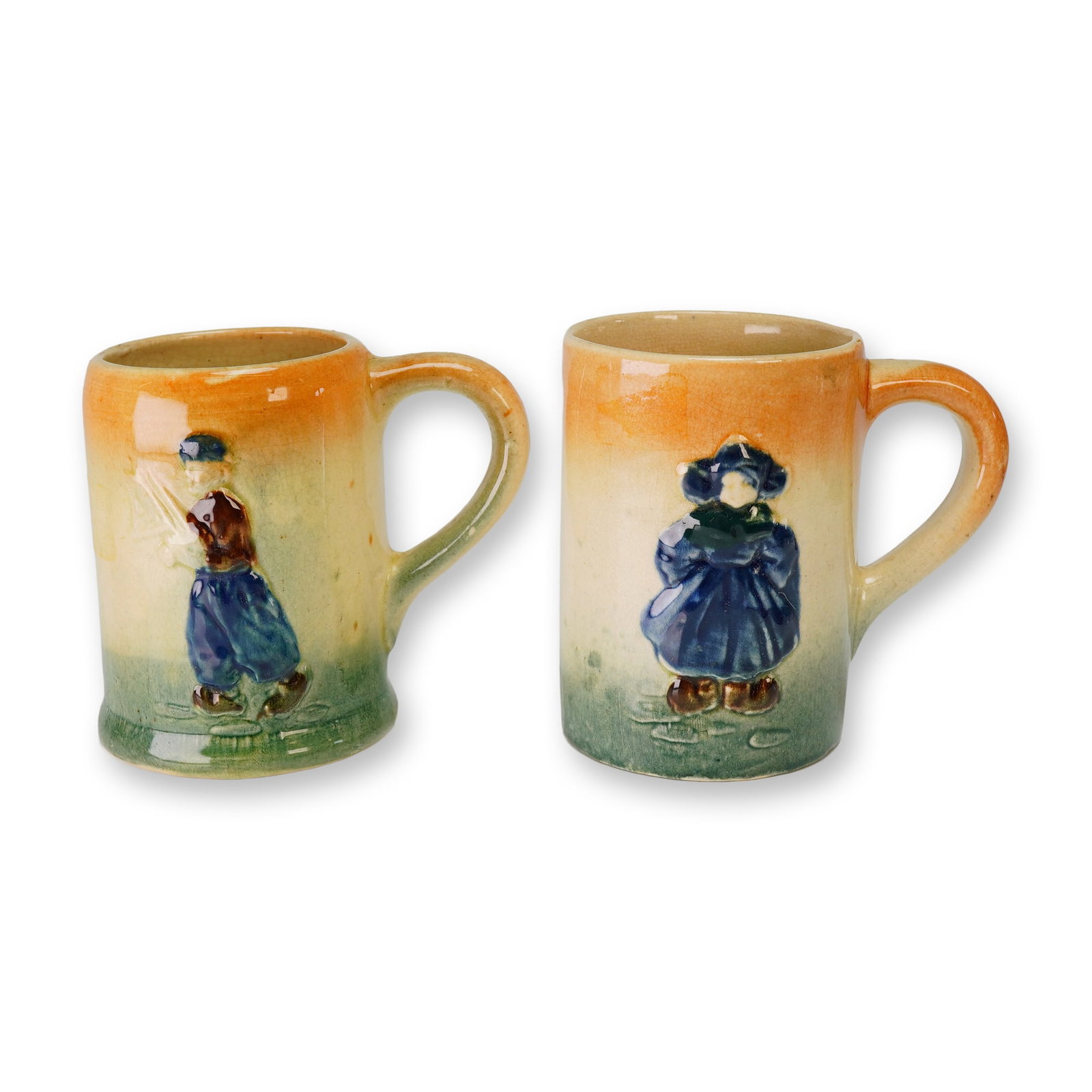 2pc Roseville Pottery Holland Ware Mugs: A pair of Roseville Holland mugs crafted from glazed stoneware. Each vessel features a raised relief figure of a child in traditional Dutch attire against a three-tone gradient background of orange, c
