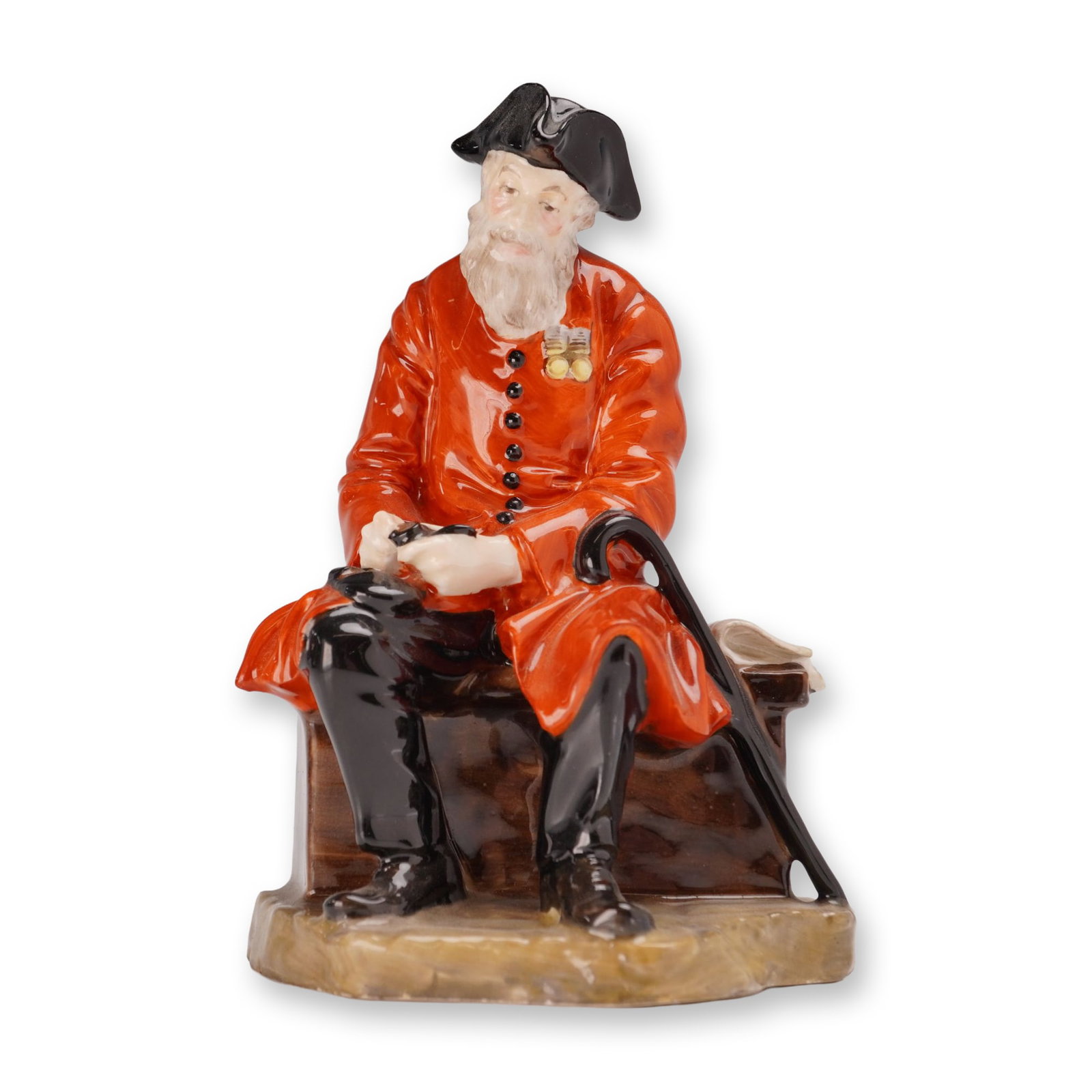 Royal Doulton Porcelain Figurine, Chelsea Pensioner HN689: Designed by Leslie Harradine. The figure is depicted seated on a brown wood-grained bench, wearing a scarlet coat with military medals, black breeches, and a black tricorn hat. He holds a pipe in his 