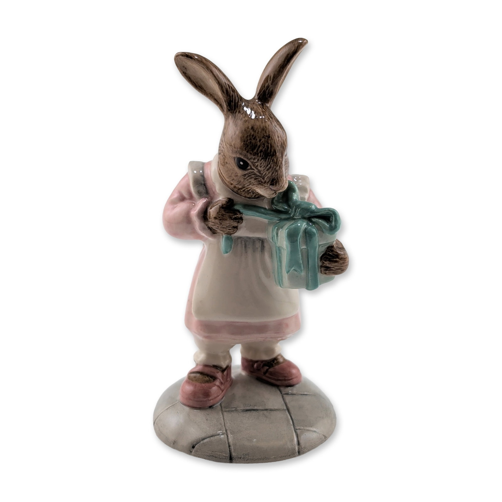 Royal Doulton Beatrix Potter Prototype Rabbit Figurine: Glazed porcelain figurine, depicting a rabbit in a pink dress and white apron carrying a gift box wrapped in blue with a teal bow. The figure is set upon a circular gray cobblestone base. The undersid