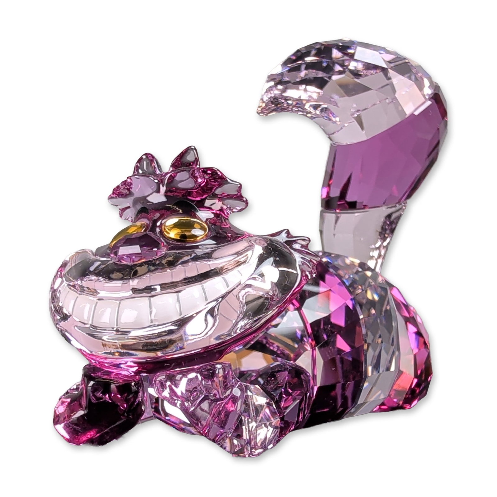 Swarovski Crystal Disney Cheshire Cat Figurine: Swarovski faceted lead crystal figurine depicting the Cheshire Cat from Disney's Alice in Wonderland. The piece is rendered in variegated shades of fuchsia, amethyst, and light pink crystal, with ambe