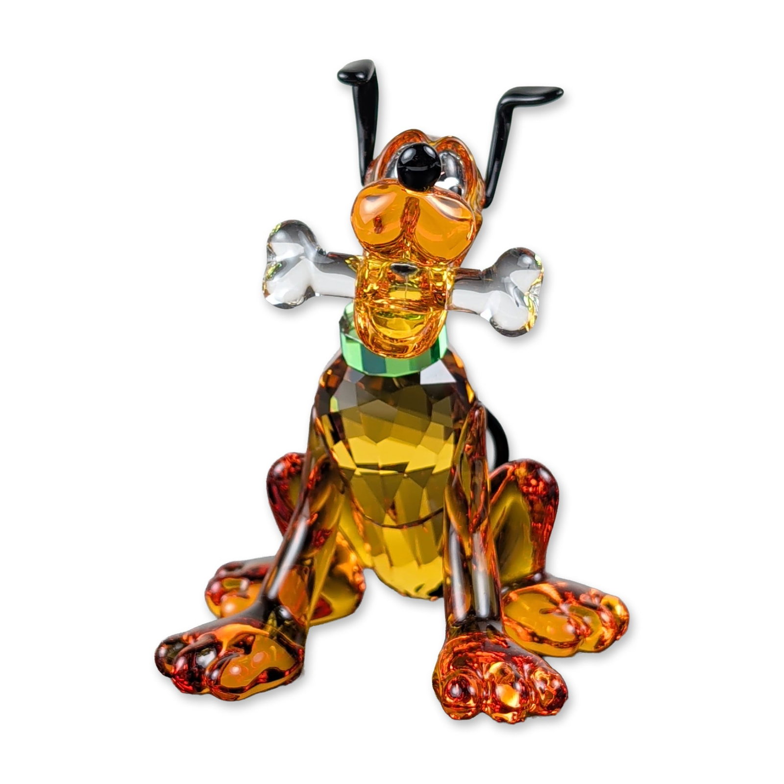 Swarovski Crystal Disney Pluto with Bone Figurine: Swarovski. Figurine of the Disney character Pluto in faceted amber lead crystal, featuring a green collar and reddish-orange highlights on the paws. Depicted in a seated position holding a frosted cle