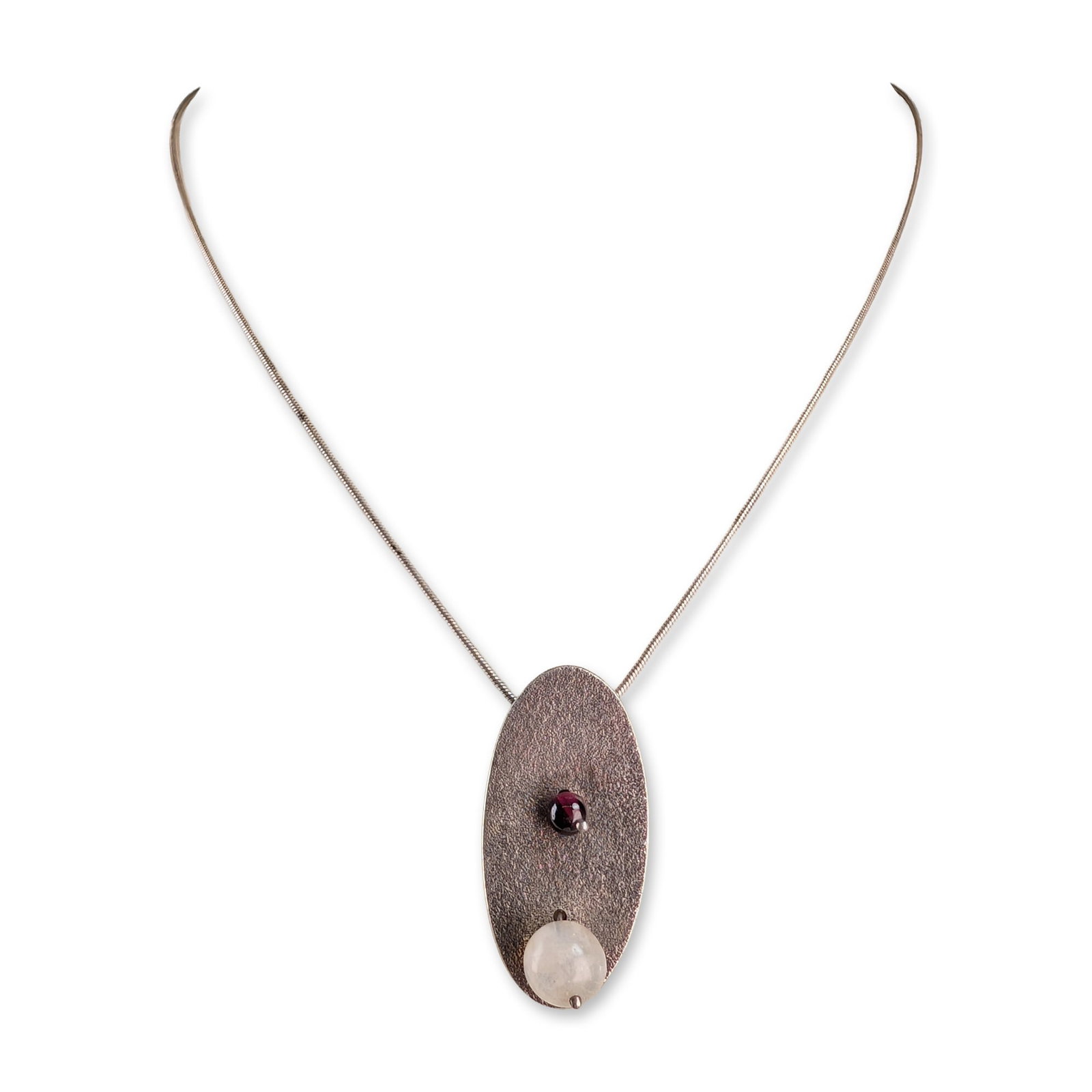 Modernist Sterling Silver Garnet Moonstone Necklace: Modernist pendant necklace in sterling silver featuring an elongated oval pendant with a stippled matte finish. The front face is set with a small round garnet cabochon and a larger translucent white 