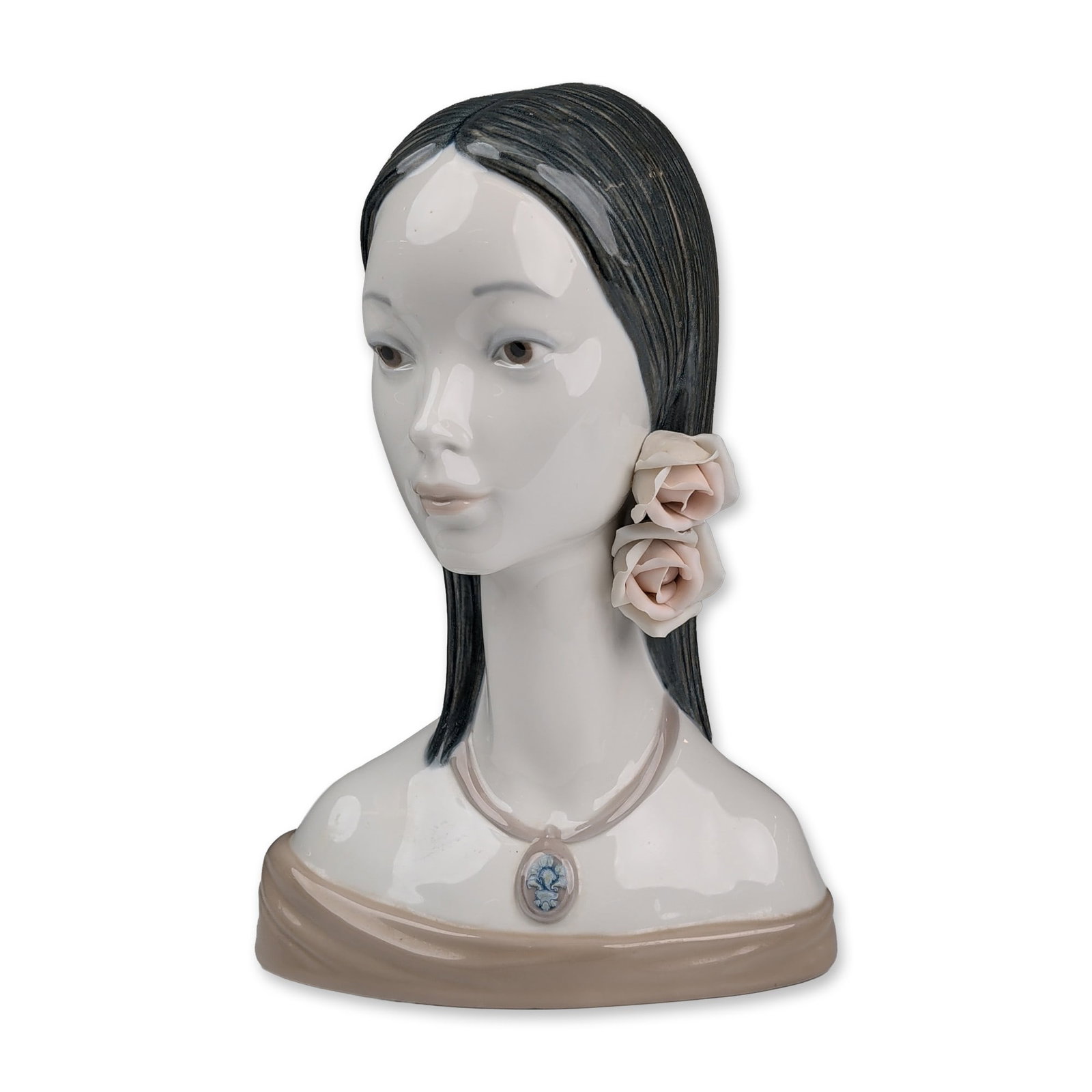 Lladro Porcelain Bust, Maja Head 1004668: Glazed porcelain bust titled La Maja, designed by Juan Huerta. The figure depicts a young woman with a downward gaze, featuring dark hair with a linear textured finish and two hand-applied pale pink p