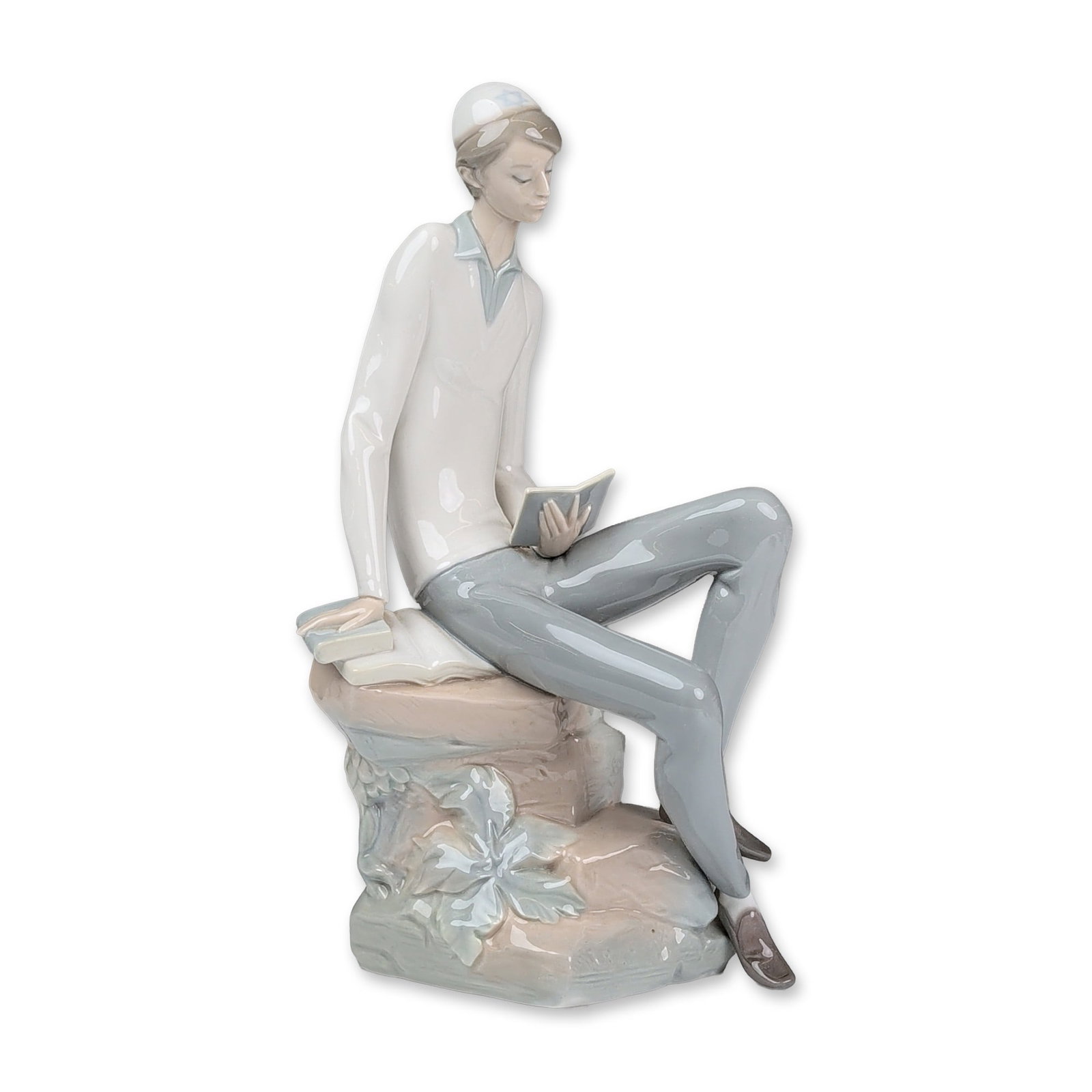 Lladro Glazed Porcelain Figurine, Hebrew Student 01004684: Figurine depicting a young man studying. The youth is seated on a textured rock-form base featuring a large relief leaf decoration. He is modeled wearing a white cap designed with Star of David, a whi