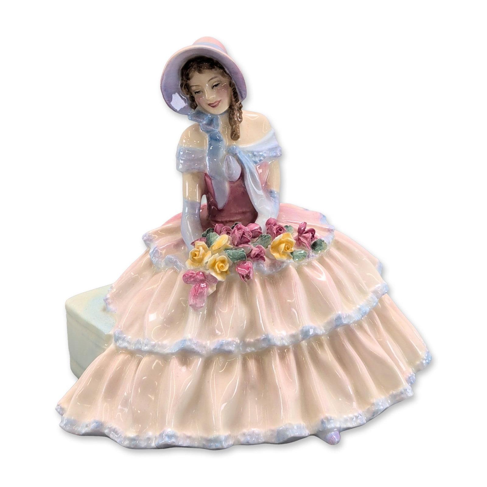 Royal Doulton Day Dreams Porcelain Figurine: Royal Doulton. Glazed bone china figurine titled Day Dreams, featuring a seated woman in a multi-tiered pink and blue gown with a lavender hat, holding a rose bouquet. The underside bears a printed ma