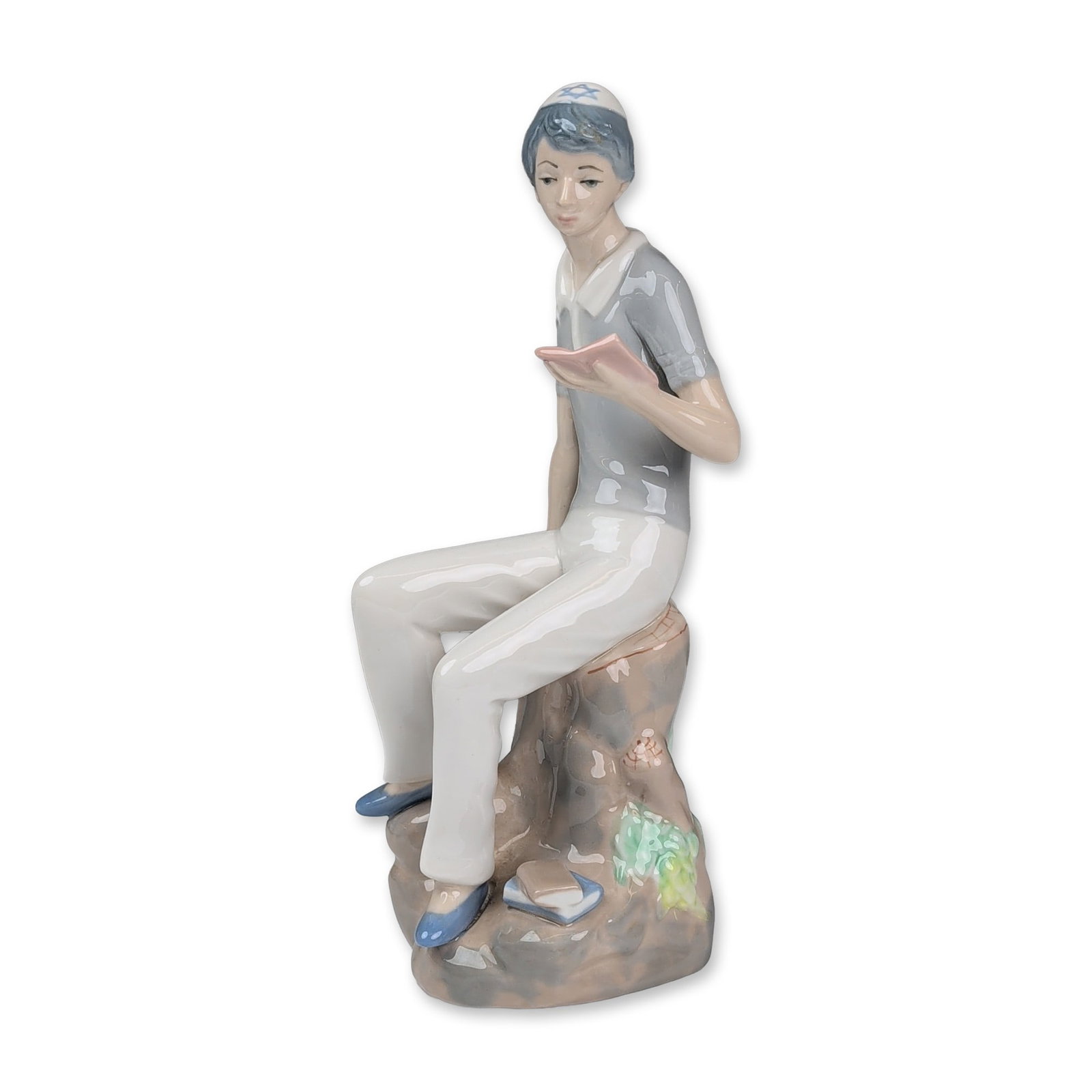 Casades Spanish Porcelain Figurine, Hebrew Student Reading: Glazed porcelain figurine depicting a young Hebrew student seated on a rocky outcrop reading a book. The figure is modeled wearing a blue and white kippah with a Star of David, a grey short-sleeved sh