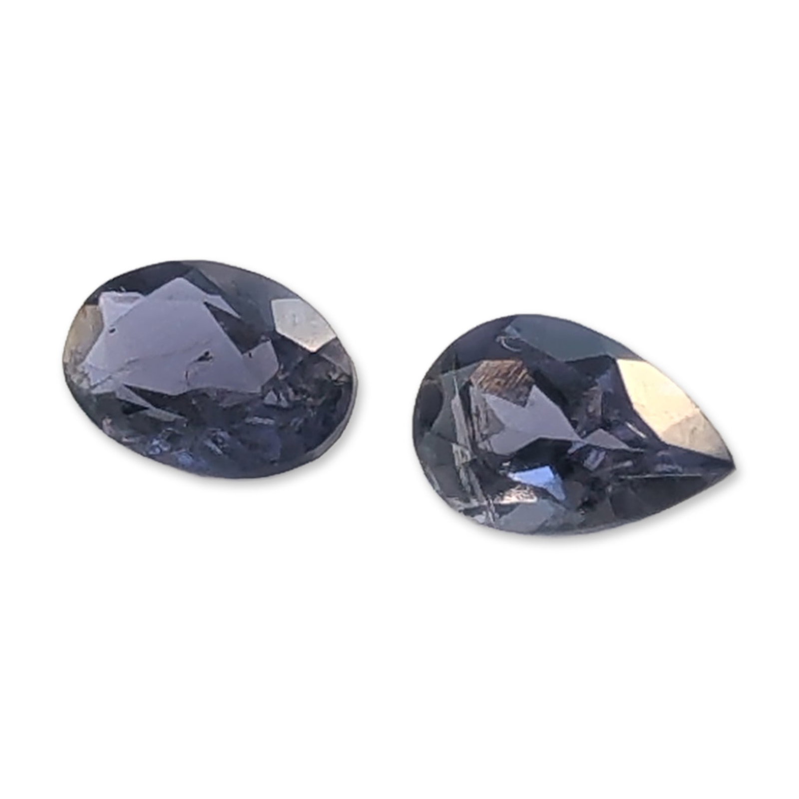 Pair of Grayish-Violet Faceted Loose Iolite Gemstones: A pair of faceted loose Iolite gemstones in a medium-dark grayish-violet or steel blue hue. The set consists of one pear-cut stone and one oval-cut stone. Visible internal characteristics are present,