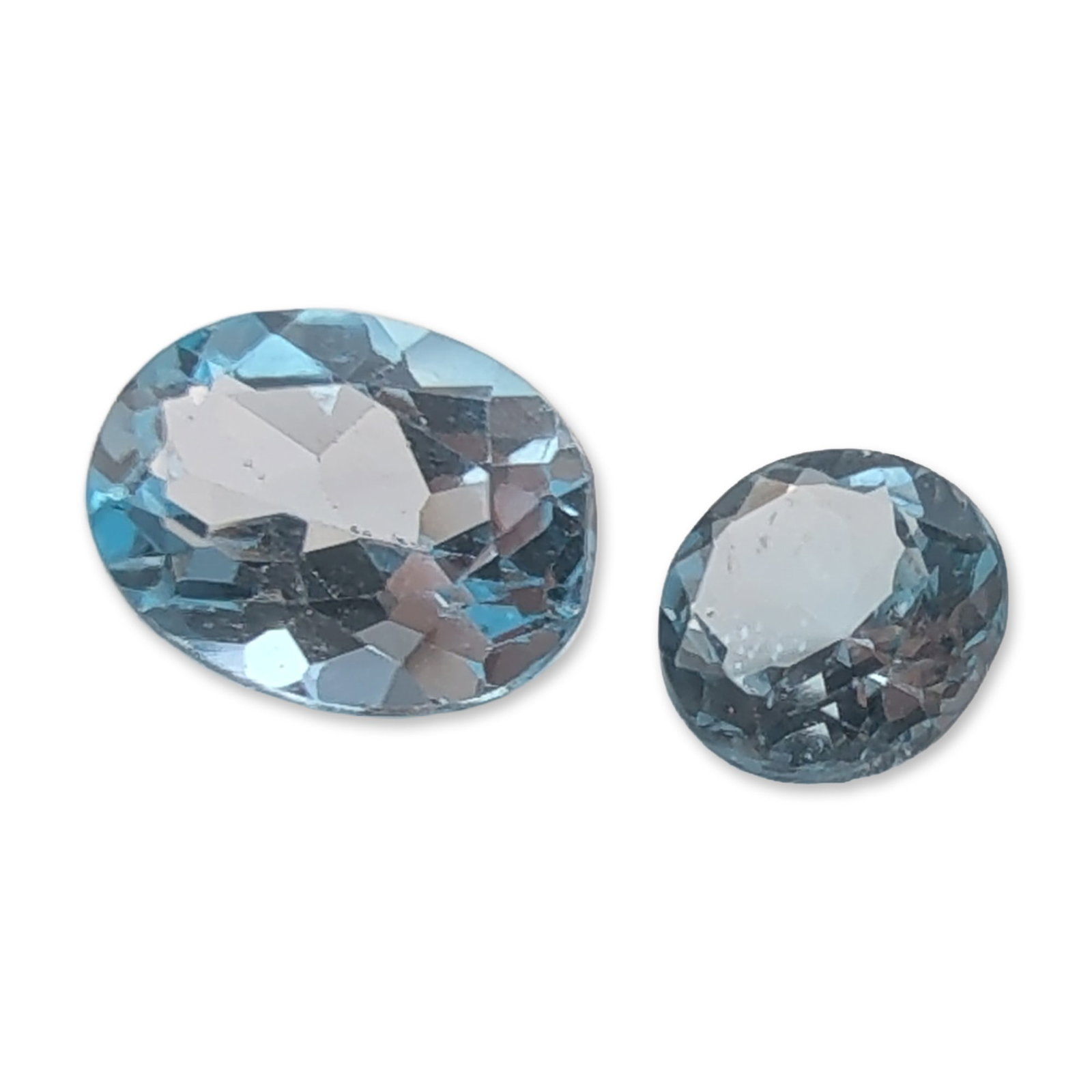 Pair of Pale Blue Faceted Topaz Gemstones: A pair of faceted light blue topaz gemstones. The group comprises one oval-cut gemstone and one round-cut gemstone, both displaying a pale sky-blue hue and good transparency. The oval stone shows cent