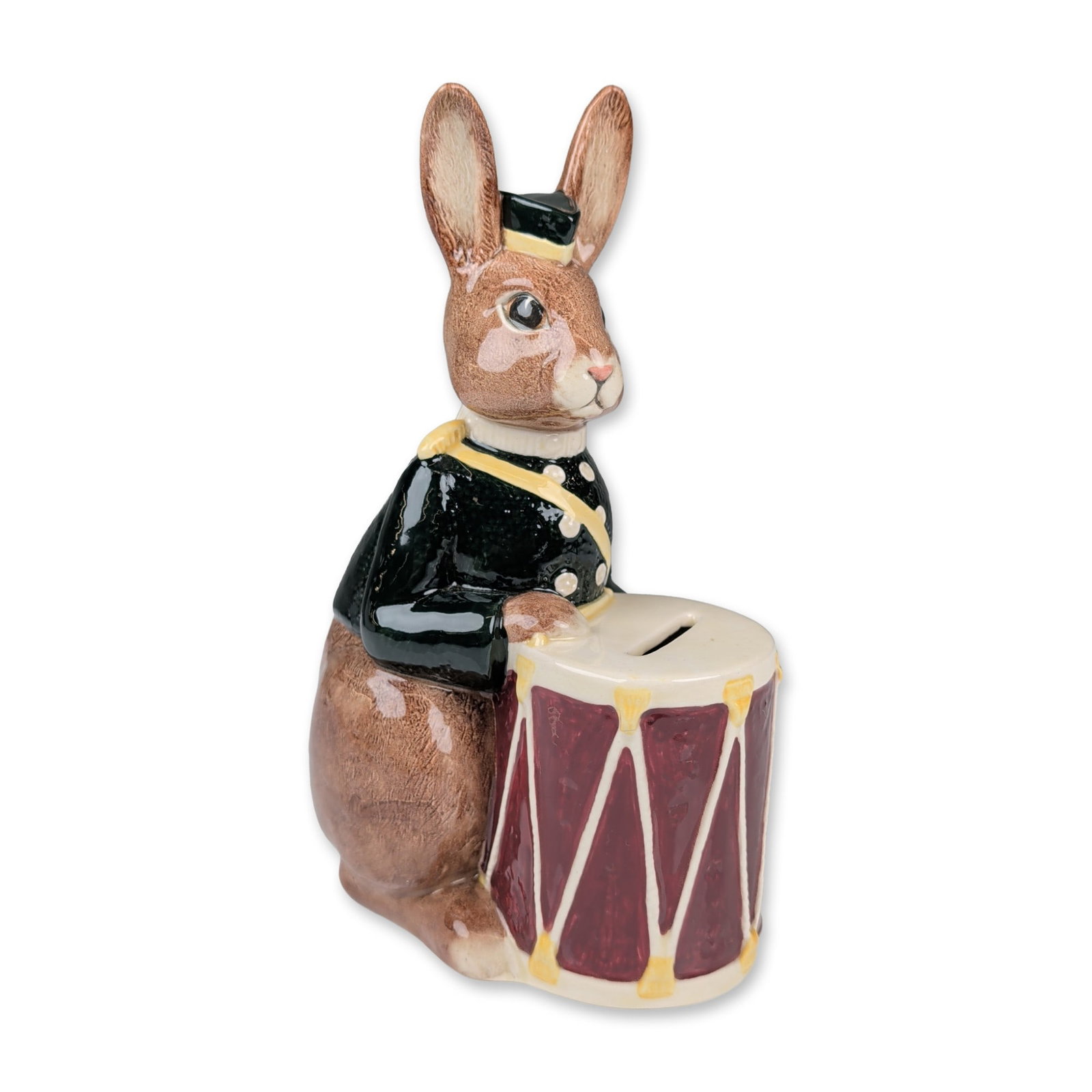 Royal Doulton Ceramic Bunnybank Drummer Bank D6615: Royal Doulton. Still bank in glazed ceramic, featuring a seated brown rabbit in military bandsman attire including a black jacket with yellow trim and a pillbox hat. The figure holds a large burgundy 