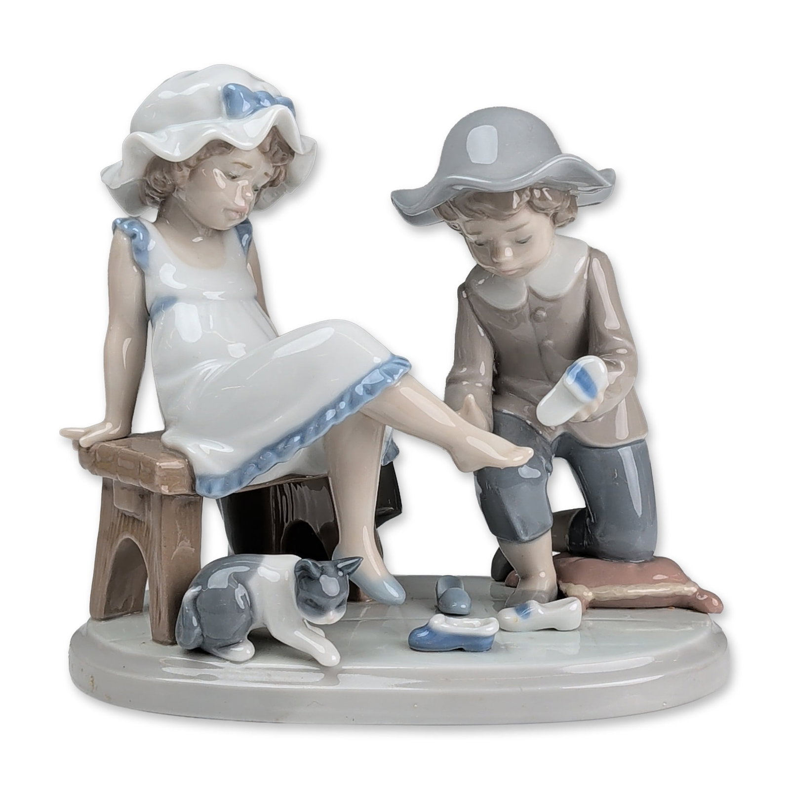 Lladro Glazed Porcelain Figurine, Try This One 01005361: The piece depicts a scene of a young boy kneeling to fit a shoe onto the foot of a girl seated on a rustic wooden bench. The girl is dressed in a white gown with ruffled blue trim and a matching volum