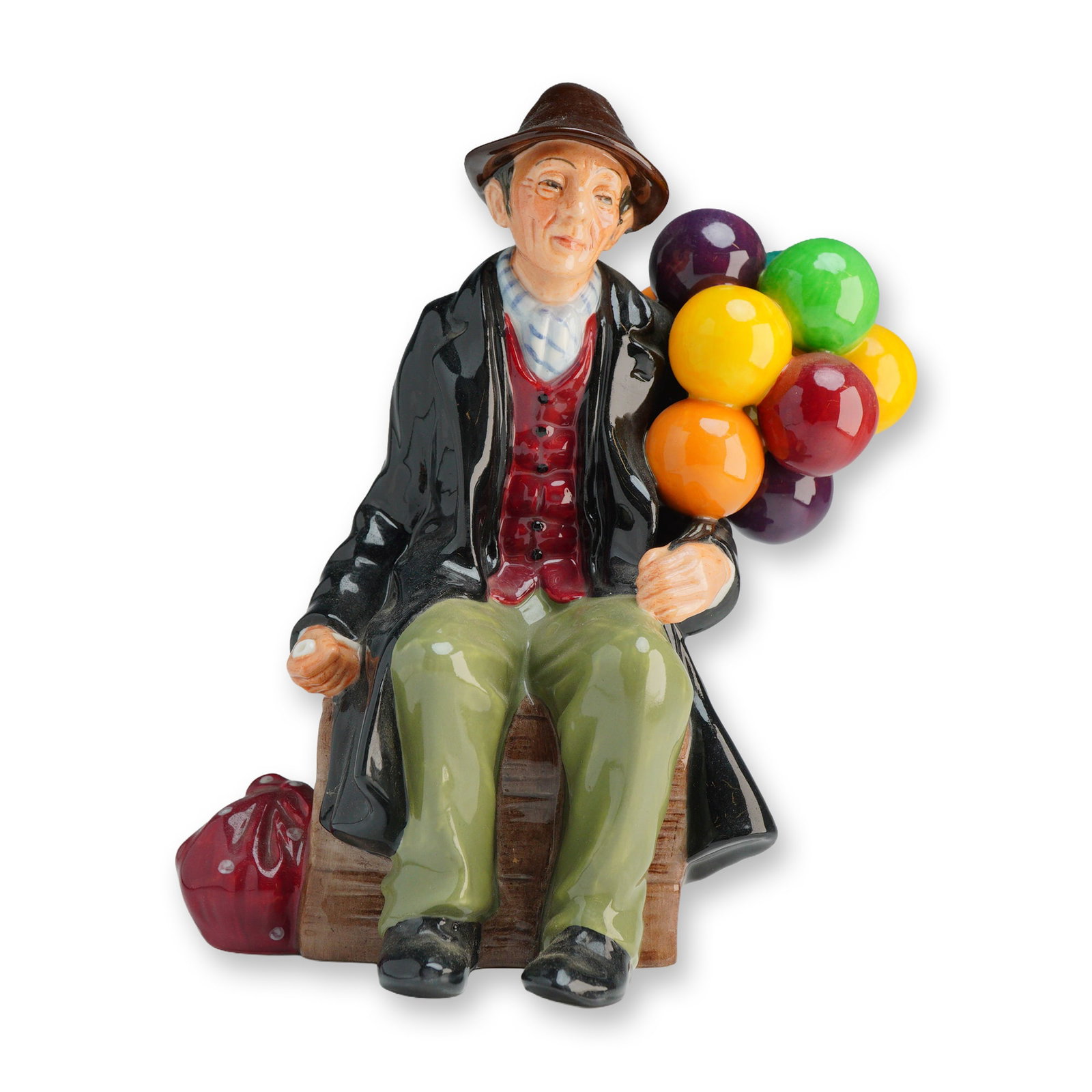 Royal Doulton Figurine The Balloon Man HN1954: This glazed porcelain figure depicts an elderly male vendor seated on a wooden-textured crate. The subject is dressed in a dark overcoat, a red waistcoat over a striped shirt, green trousers, and a br