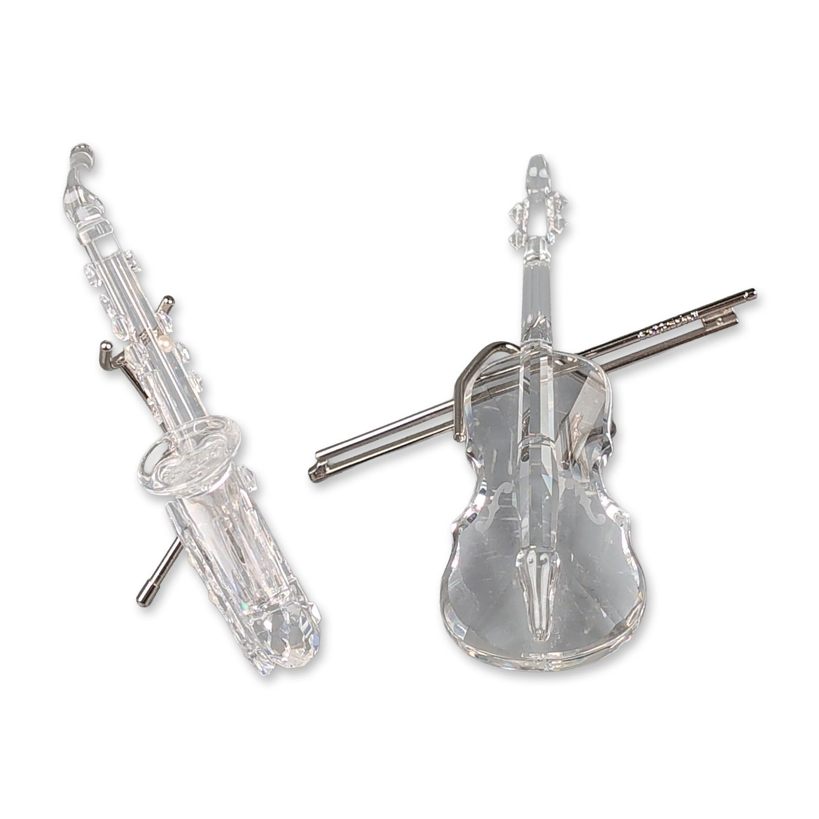 Two Swarovski Silver Crystal Musical Instrument Figurines, Violin and Saxophone: Two miniature musical instrument figurines in faceted clear lead crystal. The violin is accompanied by a rhodium-plated metal bow that functions as a stand, featuring a textured section to represent