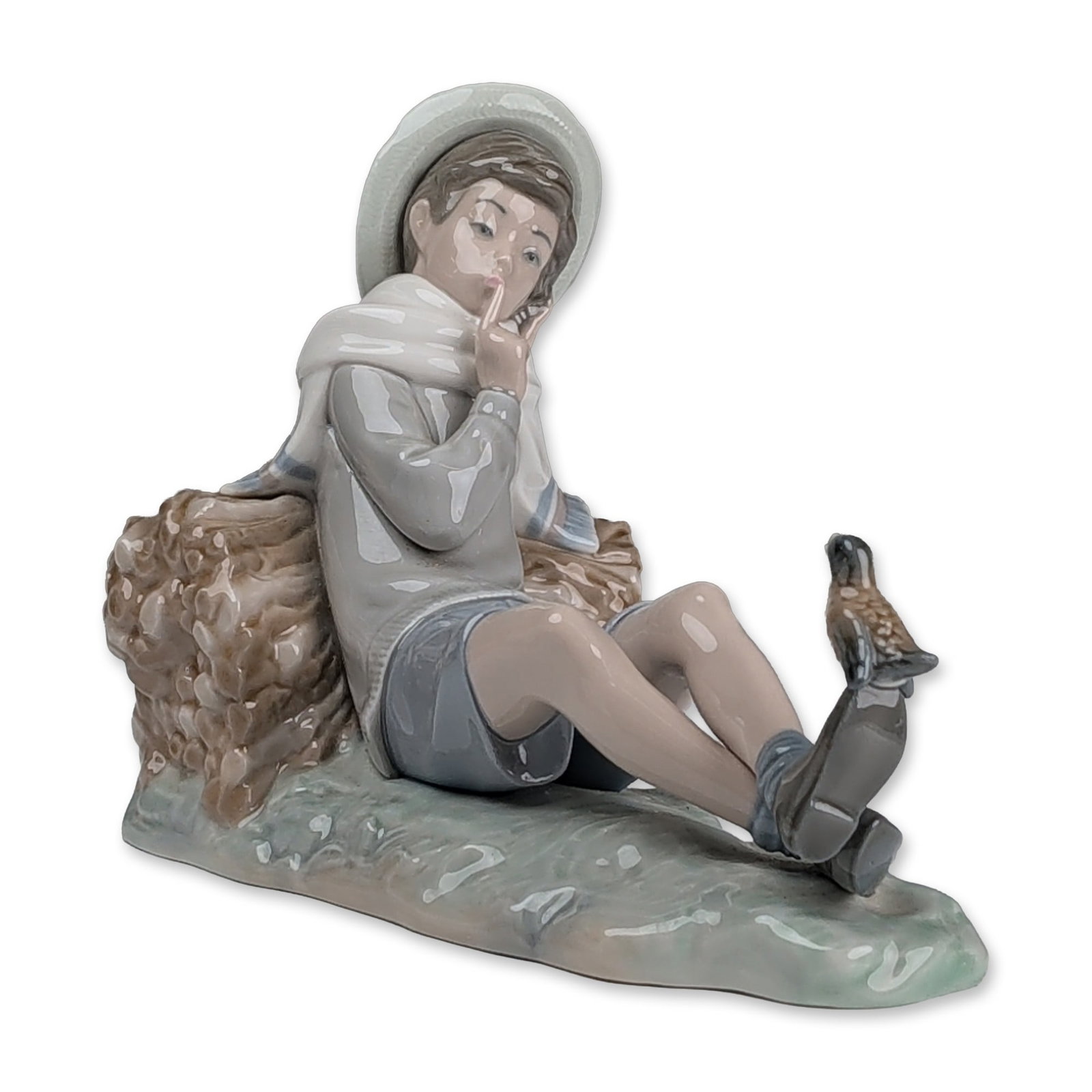 Lladro Glazed Porcelain Figurine, Shepherd with Bird 01004730: Lladro (Spain) glazed porcelain figurine designed by Vincente Martinez. The polychrome piece depicts a young boy reclining against a bound bundle of sticks, wearing a grey long-sleeved sweater, blue s