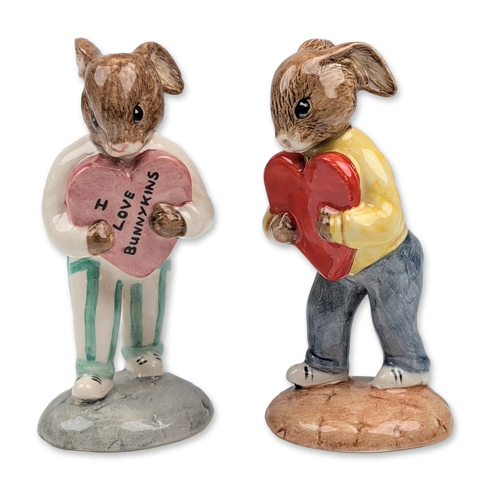 Pair of Royal Doulton Sweetheart Bunnykins Figurines: Royal Doulton. Pair of glazed ceramic figurines from the Bunnykins series, both in the Sweetheart model. The first figurine, model DB 130, depicts a rabbit in a yellow sweater and blue trousers holdin