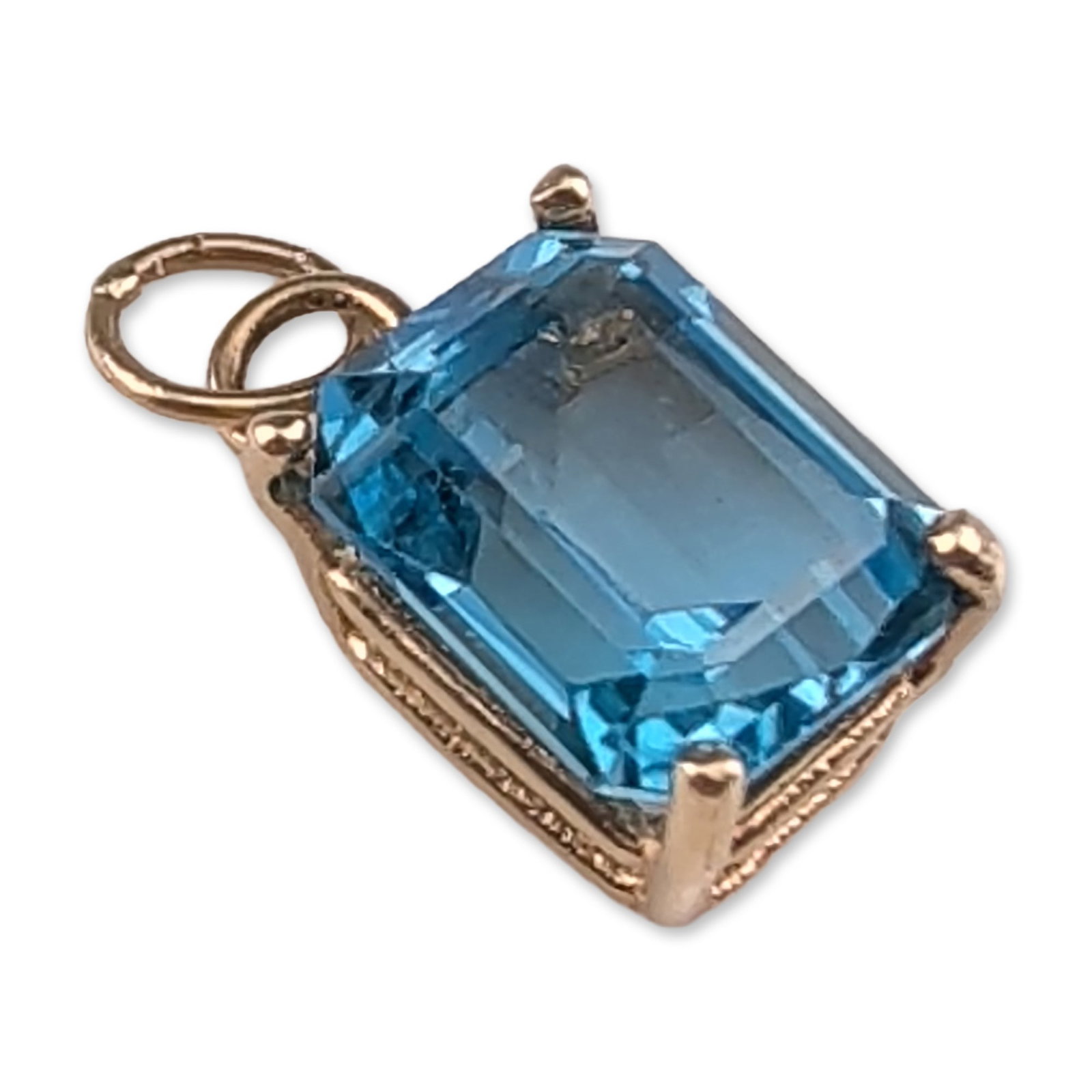 14K Yellow Gold Emerald Cut Blue Topaz Pendant Charm: Yellow gold pendant charm featuring an emerald-cut blue topaz. The stone is secured in a four-prong basket setting with an open gallery. The piece is fitted with a soldered jump ring and a secondary s