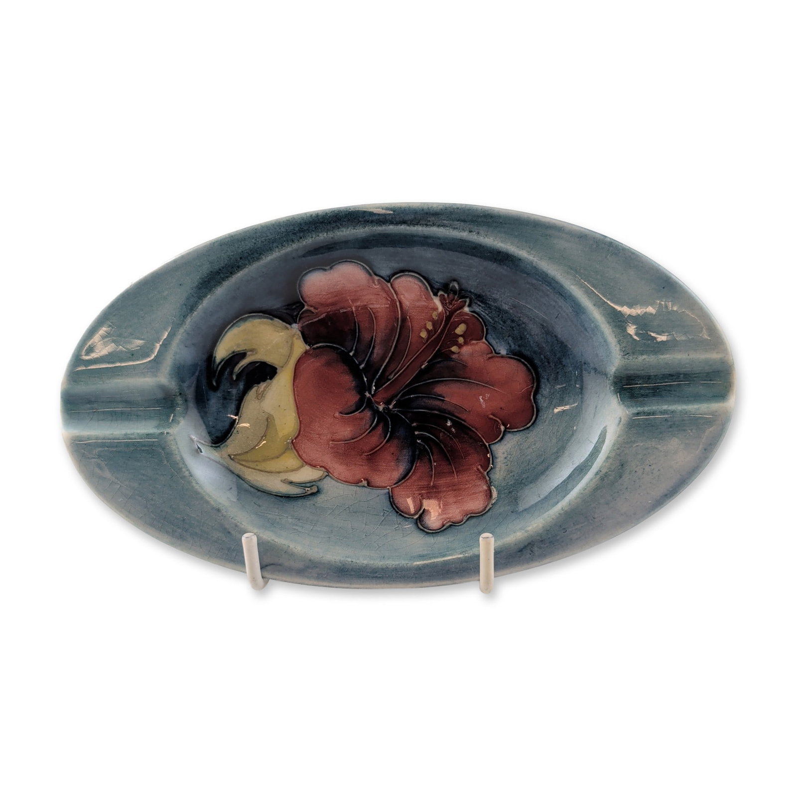 Moorcroft Hibiscus Pattern Pottery Ashtray: Moorcroft pottery oval ashtray in the Hibiscus pattern, featuring tubelined floral decoration with a red flower and yellow foliage set against a blue-grey wash ground. The form includes two integrated