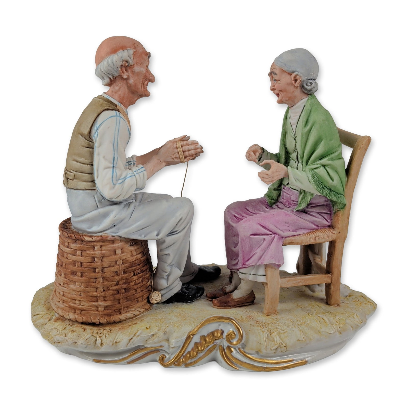Italian Porcelain Elderly Couple Figurine: Italian hand-painted porcelain genre group depicting an elderly couple and a dog. The scene features a man seated on a wicker-textured basket winding a length of cord and a woman seated in a ladder-ba