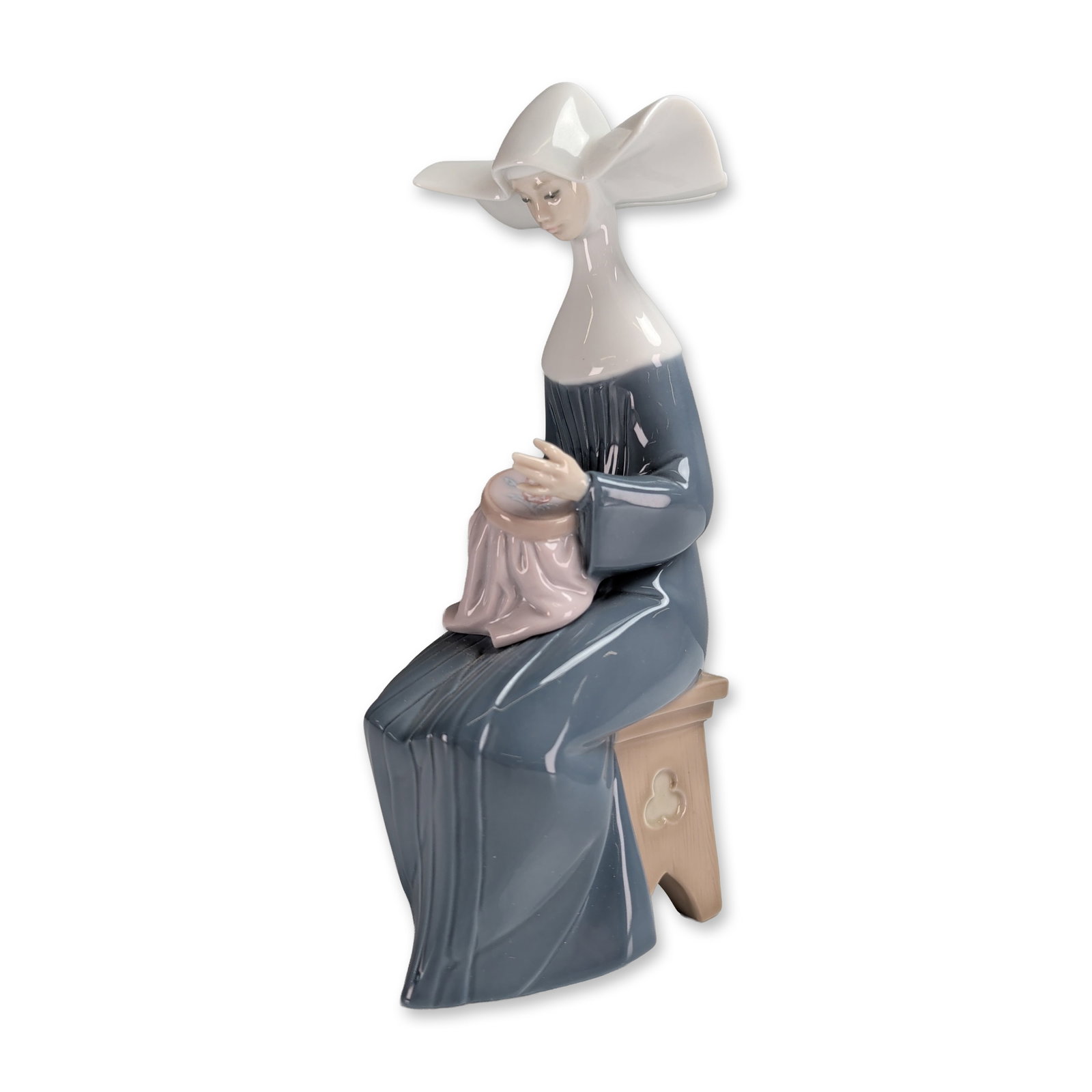 Lladro Glazed Porcelain Figurine, Time to Sew - Blue 1005501: This piece depicts a nun in a muted blue habit and white cornette hat, seated on a stool and performing needlework on an embroidery hoop. The base features the Lladro backstamp. Includes the original 