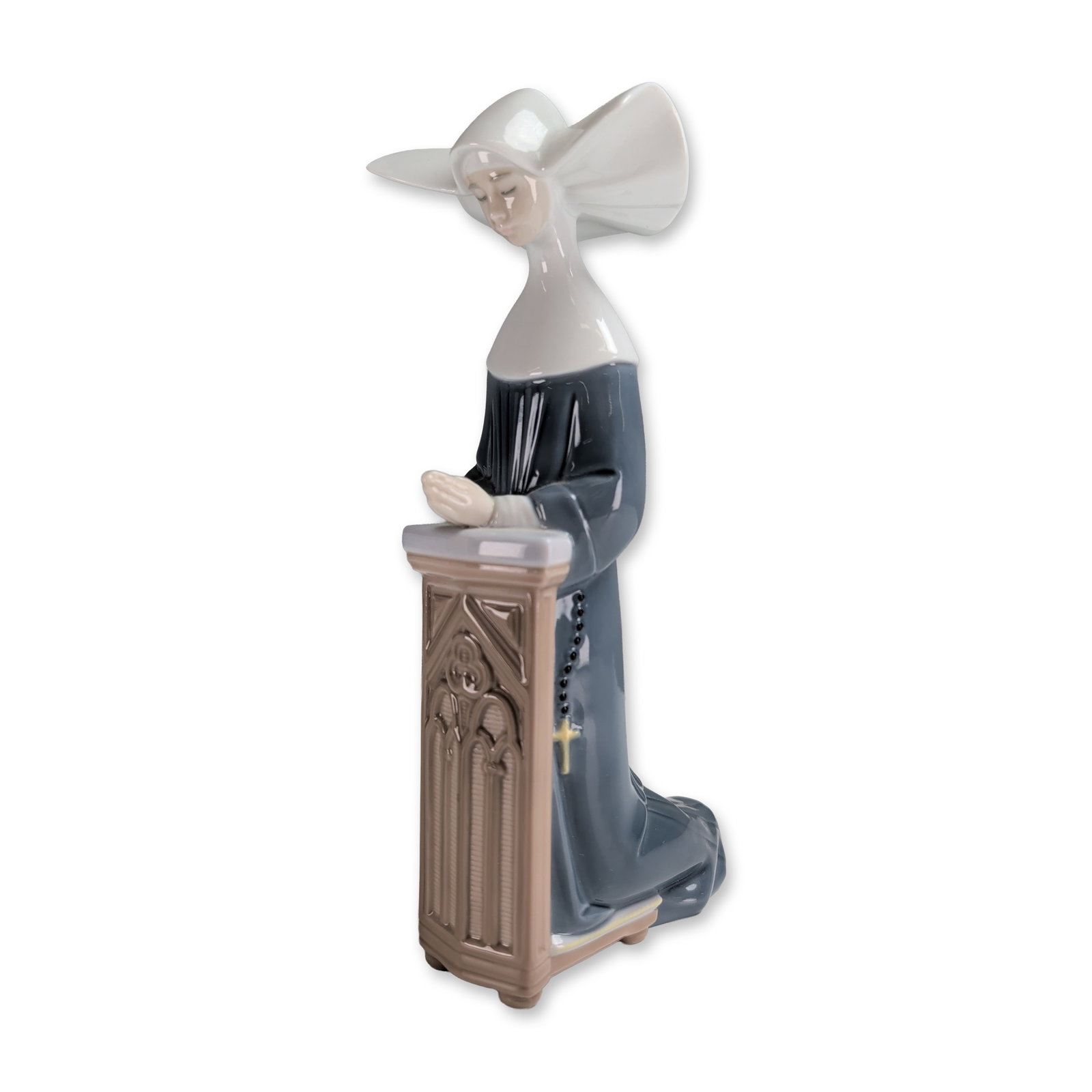 Lladro Glazed Porcelain Figurine, Meditation - Blue 01005502: Depicting a nun in a grey habit and white winged cornette kneeling in prayer at a tan-colored prie-dieu. The figure holds a rosary with black beads and a gold-toned cross. The underside bears the Llad