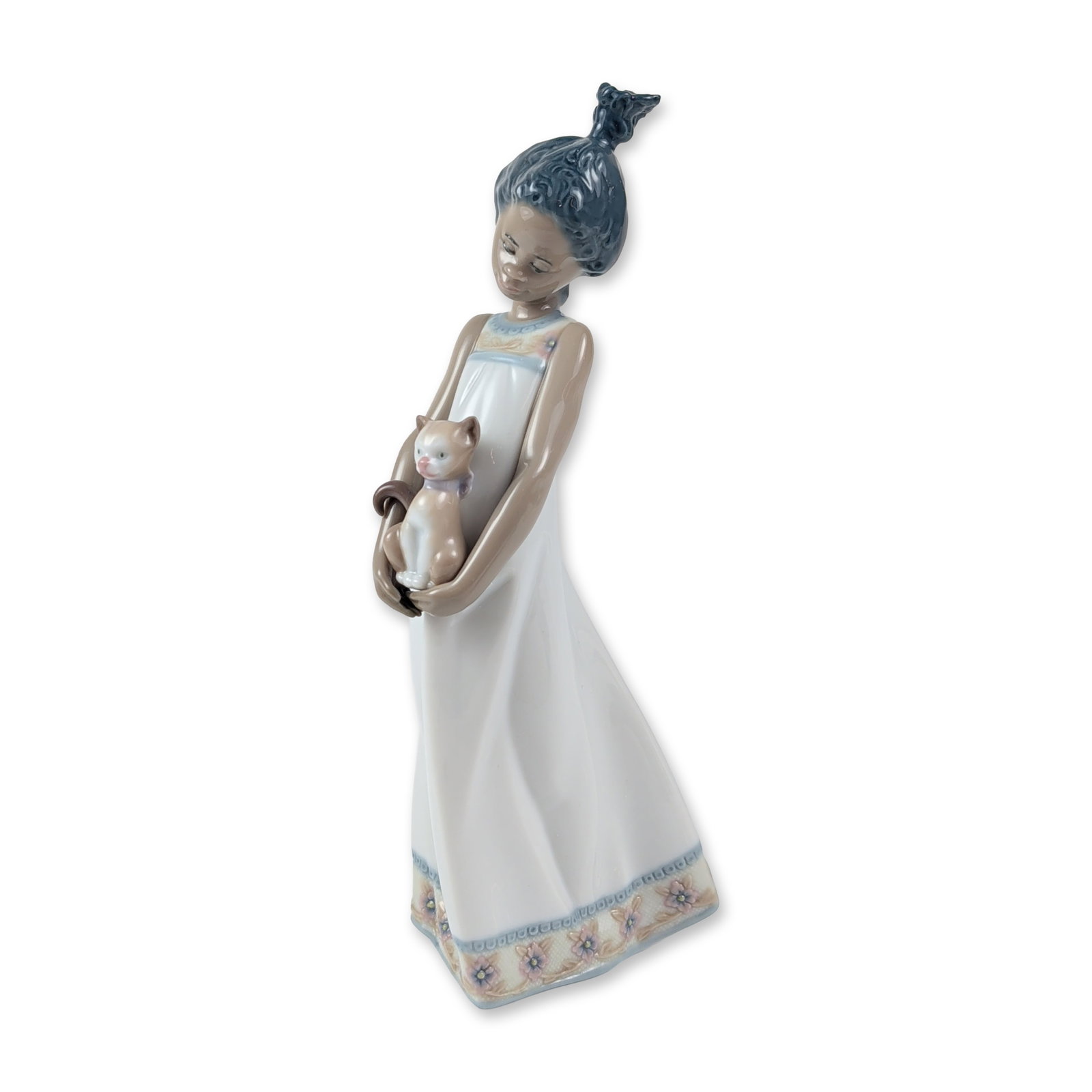 Lladro Porcelain Figurine, Close to My Heart 1005603: Depicting a young girl with dark skin tones and dark hair in a top-knot bun, wearing a white gown with floral borders at the neck and hem, cradling a kitten. Base bears Lladro backstamp. Accompanied b
