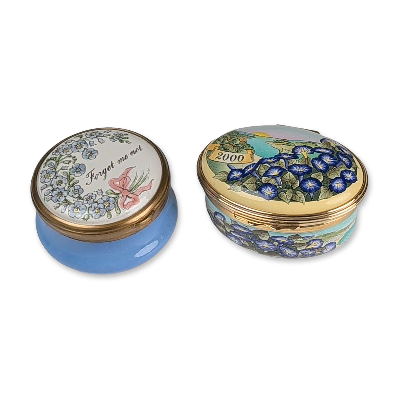 2pc Halcyon Days English Enamel Trinket Boxes: Two enamel on copper trinket boxes with gilded metal mounts. The circular box features a light blue base and a lid decorated with forget-me-not flowers and a pink ribbon bow inscribed Forget me not. A