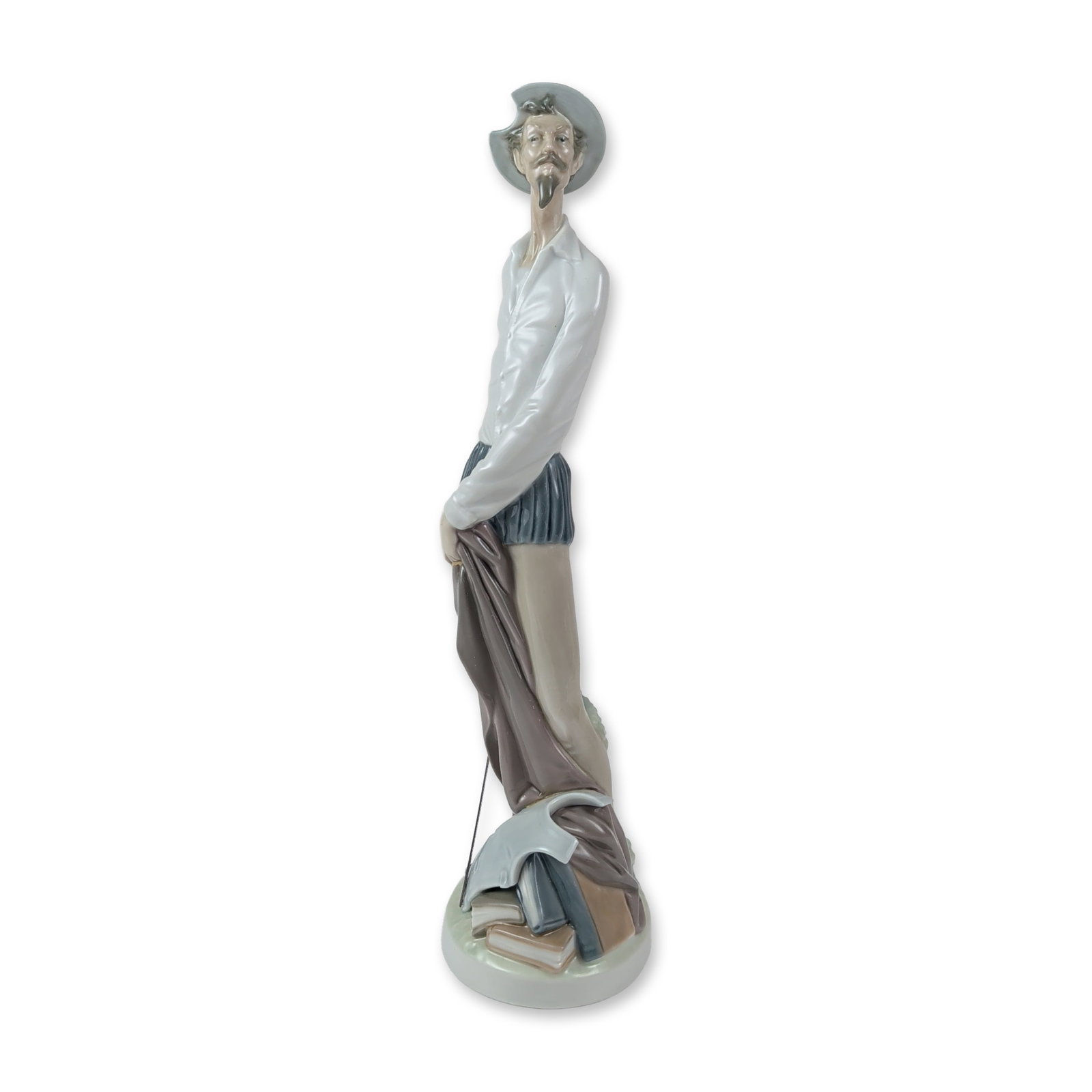 Lladro Porcelain Figurine, Quixote Standing Up 1004854: Depicting Don Quixote in a standing pose, holding a cloak and leaning over a stack of books and a shield. The figure is rendered in an elongated, stylized form with a palette of gray, blue, tan, and w
