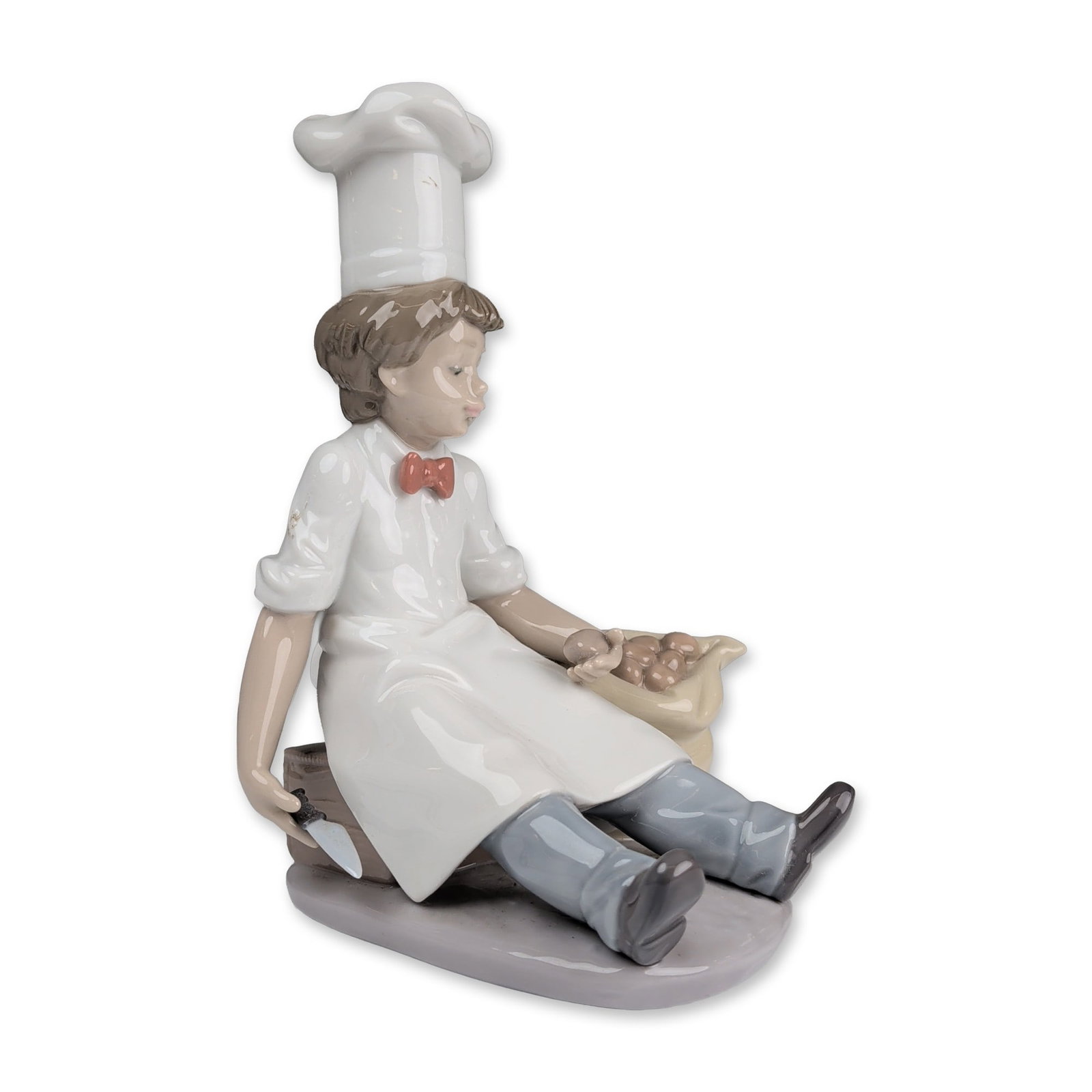 Lladro Glazed Porcelain Figurine, Chefs Apprentice 01006233: A Lladro glazed porcelain figurine depicting a seated boy in traditional chef attire including a white toque, short-sleeved shirt with red bow tie, and white apron. The figure is posed holding a parin