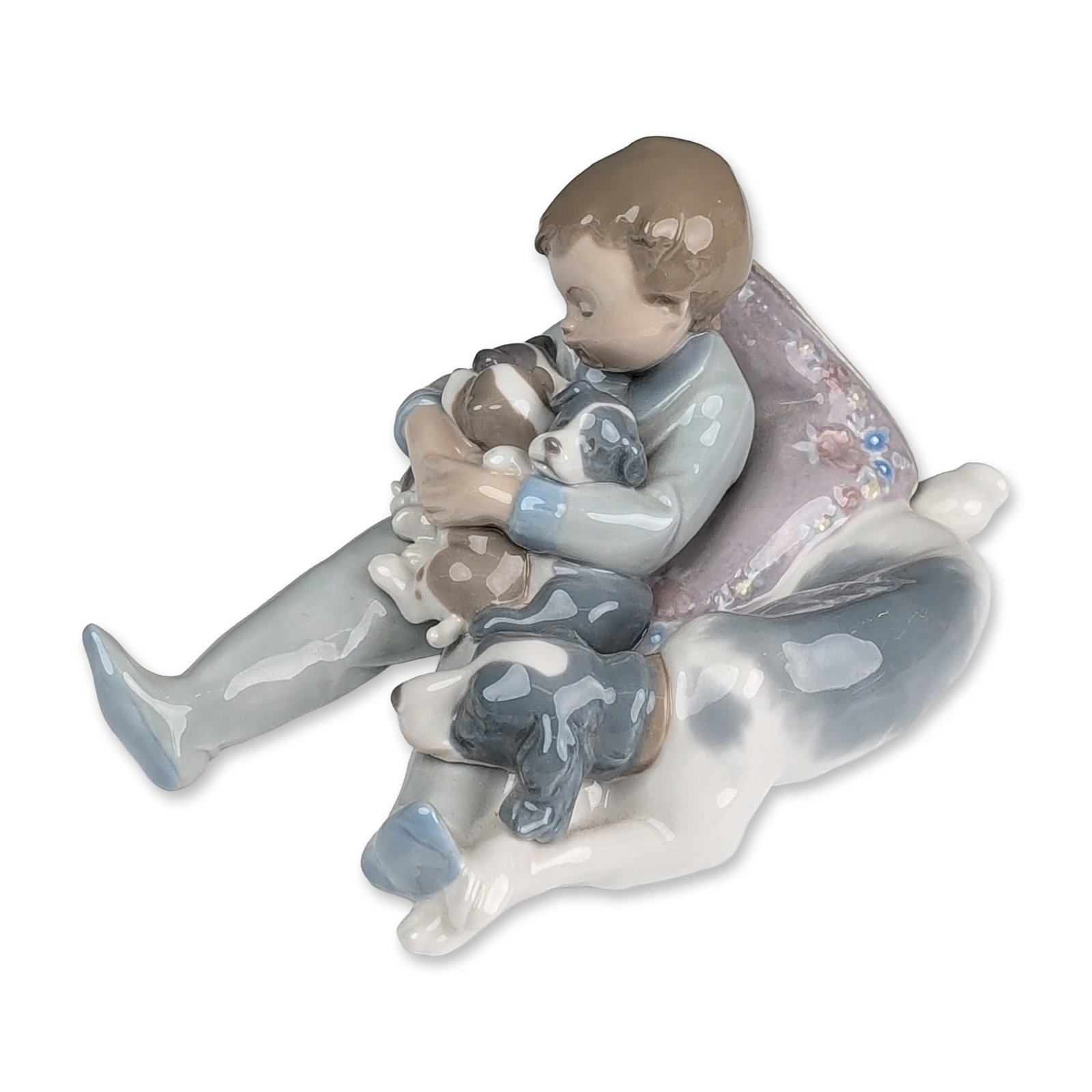 Lladro Spanish Porcelain Figurine, Sweet Dreams 01001535: A Lladro Spanish glazed porcelain figurine, model 01001535, titled Sweet Dreams, depicting a young boy in blue pajamas reclining on a lilac pillow with relief floral details while holding three puppie
