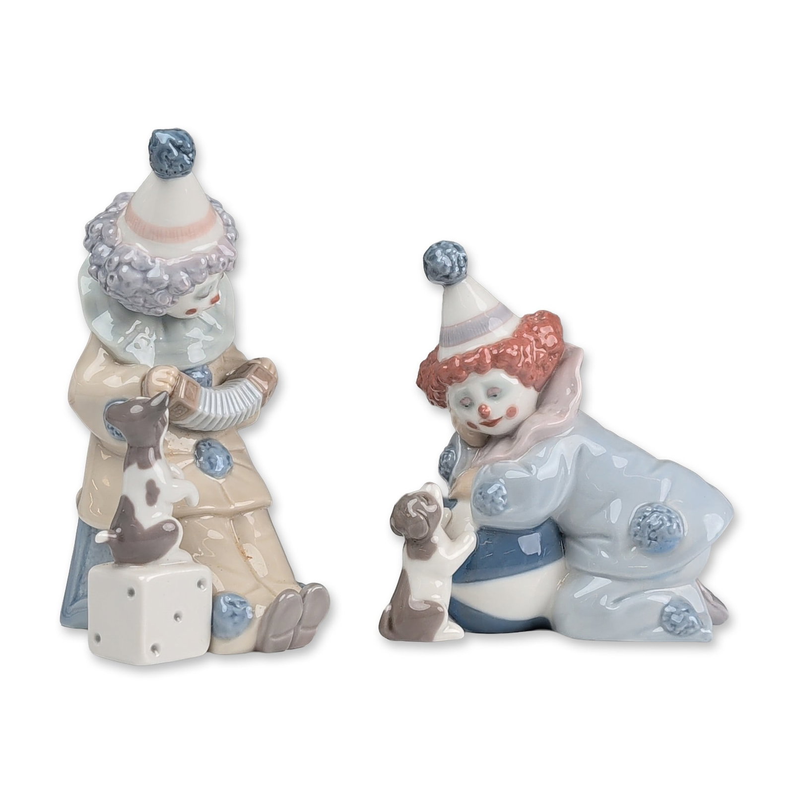 2pc Lladro Porcelain Pierrot Figurines, with Puppy 01005278 & with Concertina 01005279: Glazed porcelain figurines manufactured in Spain, each depicting a Pierrot clown. One figurine features a seated clown in a beige suit with blue pom-poms playing a concertina for a spotted dog perched