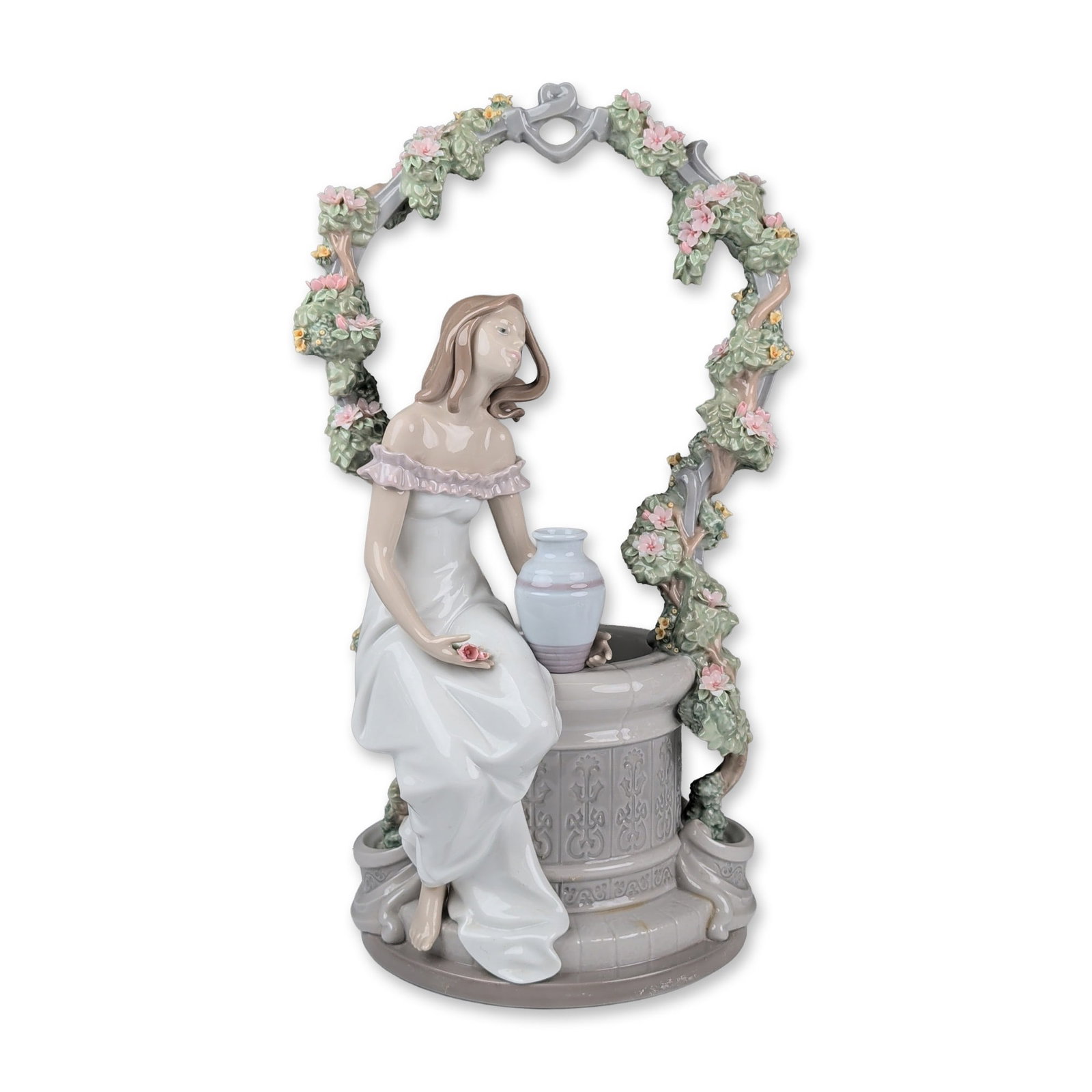 Lladro Porcelain Sculpture, A Wish For Love 01006562: Glazed porcelain figurine designed by Joan Coderch featuring a young woman in a white gown seated at a stone well beneath a floral trellis arch. The piece includes hand-applied porcelain flowers in sh