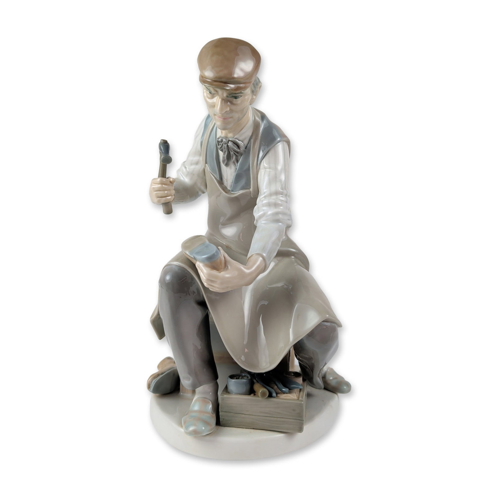 Lladro Spanish Glazed Porcelain Cobbler Figurine 1004853: Lladro Cobbler figurine, model number 1004853, in glazed porcelain. Designed by Salvador Furio, the figure depicts a seated shoemaker wearing a flat cap, pinstriped shirt, grey vest, and tan apron, ho
